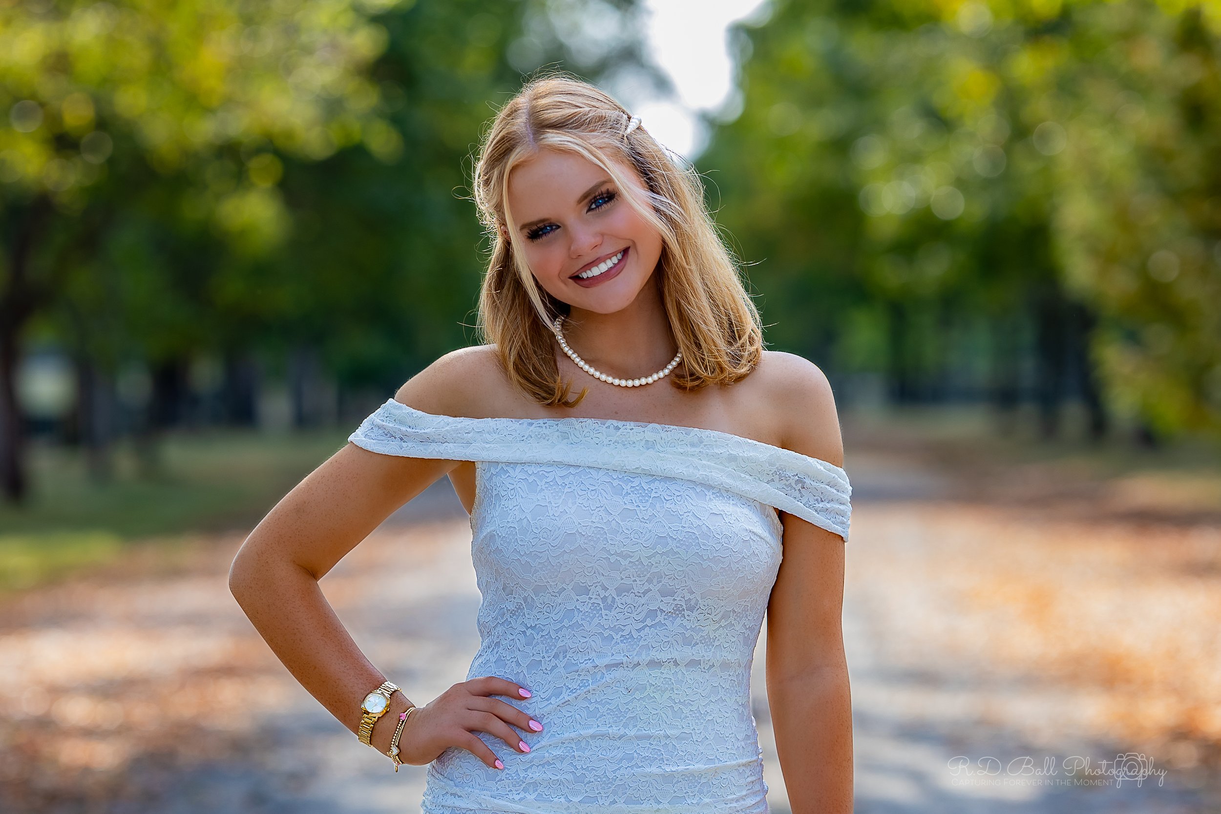 A young woman with blonde hair styled in loose waves, wearing an off-the-shoulder white lace dress, pearl necklace, gold watch, and bracelet, standing outdoors on a path in a park with trees and greenery in the background, smiling and posing with her