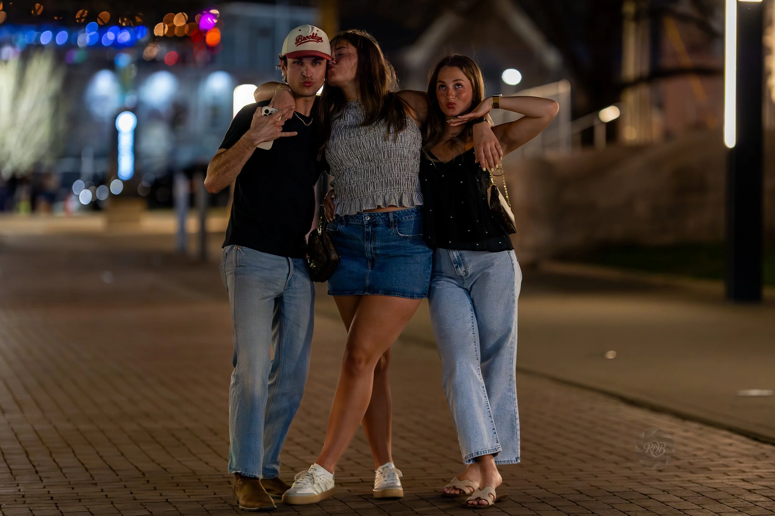 Three young adults posing together on a city sidewalk at night, with blurred colorful lights in the background. The person on the left is a young man wearing a Brooklyn baseball cap, black t-shirt, and jeans. The person in the middle is a young woman