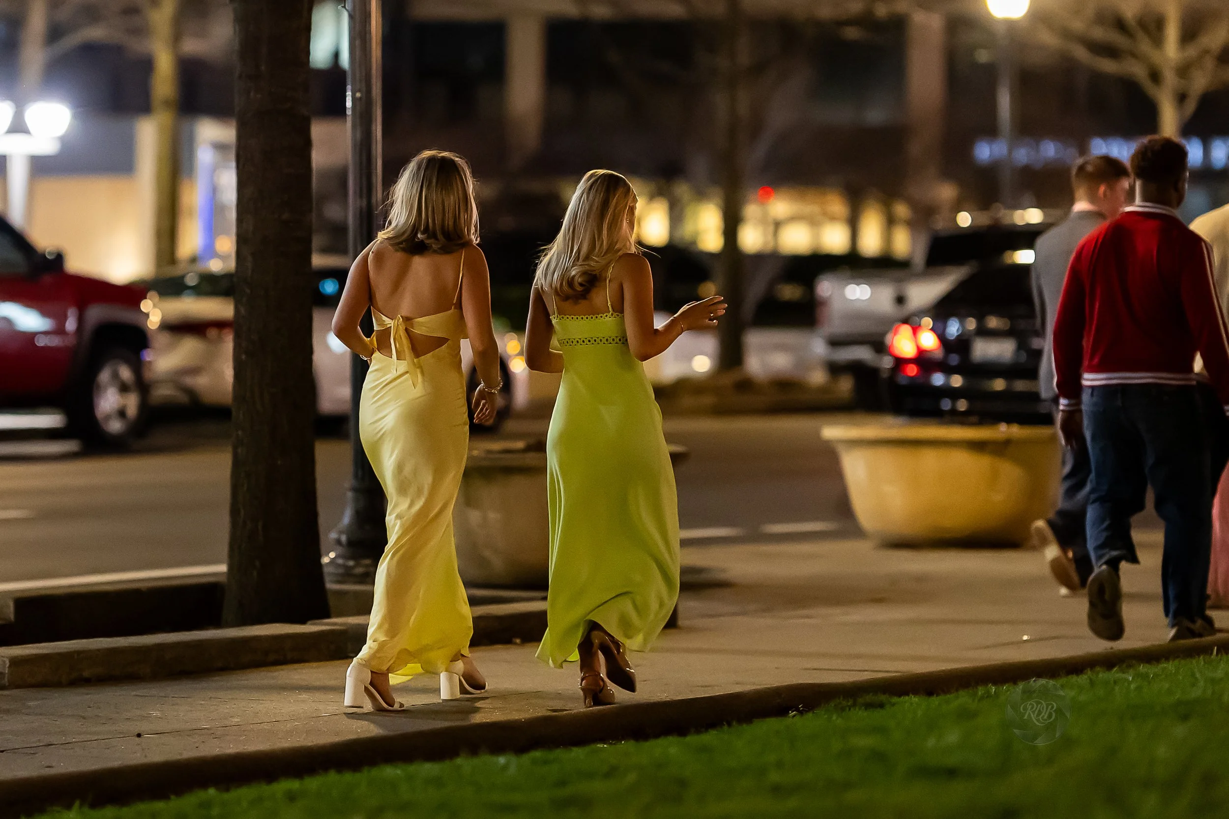 Two women in elegant yellow dresses walking on a city sidewalk at night, with parked cars and other pedestrians in the background.