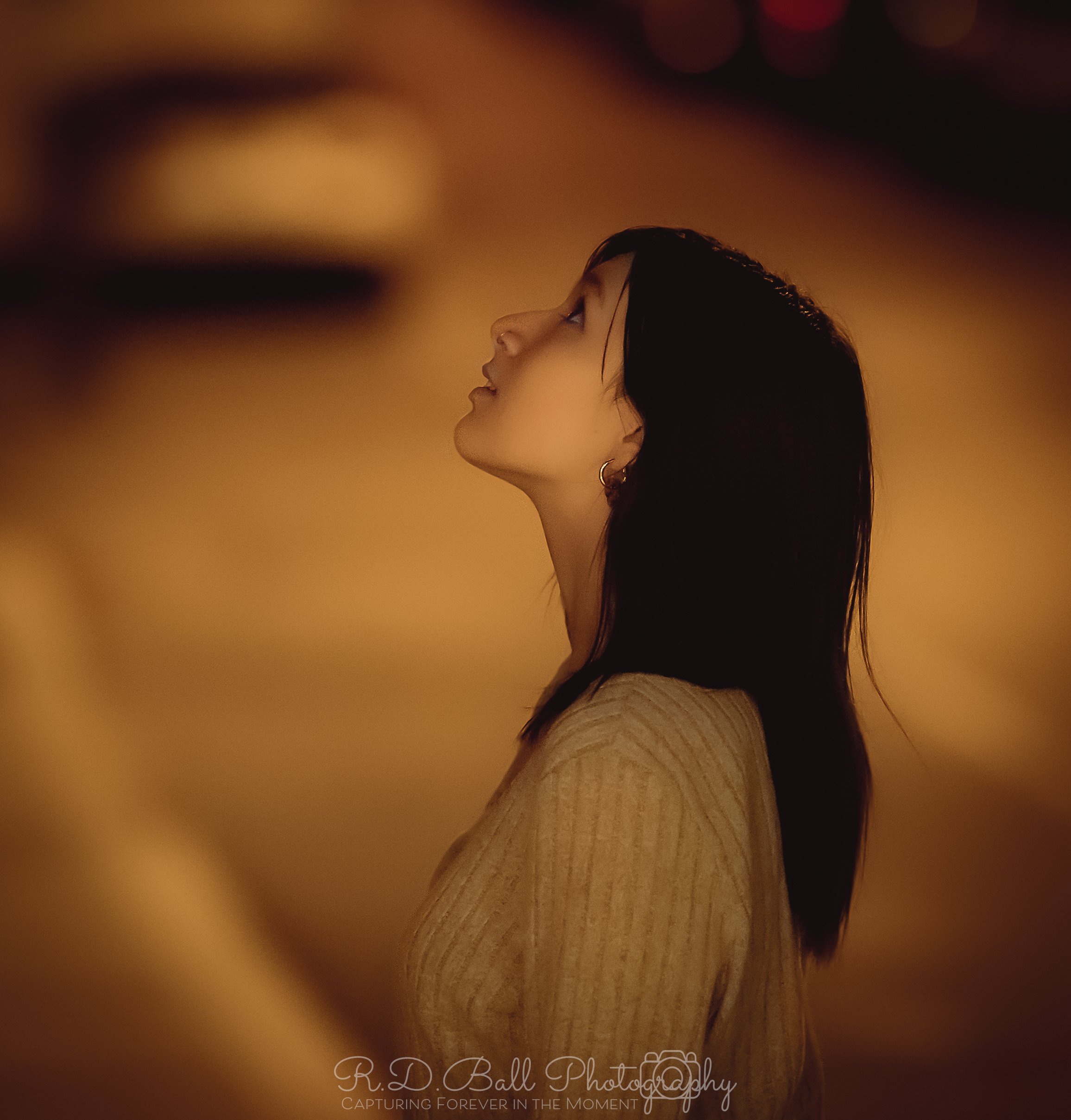 A young woman with dark hair, wearing a beige sweater, looking to the left with a calm expression, against a warm, blurred background.