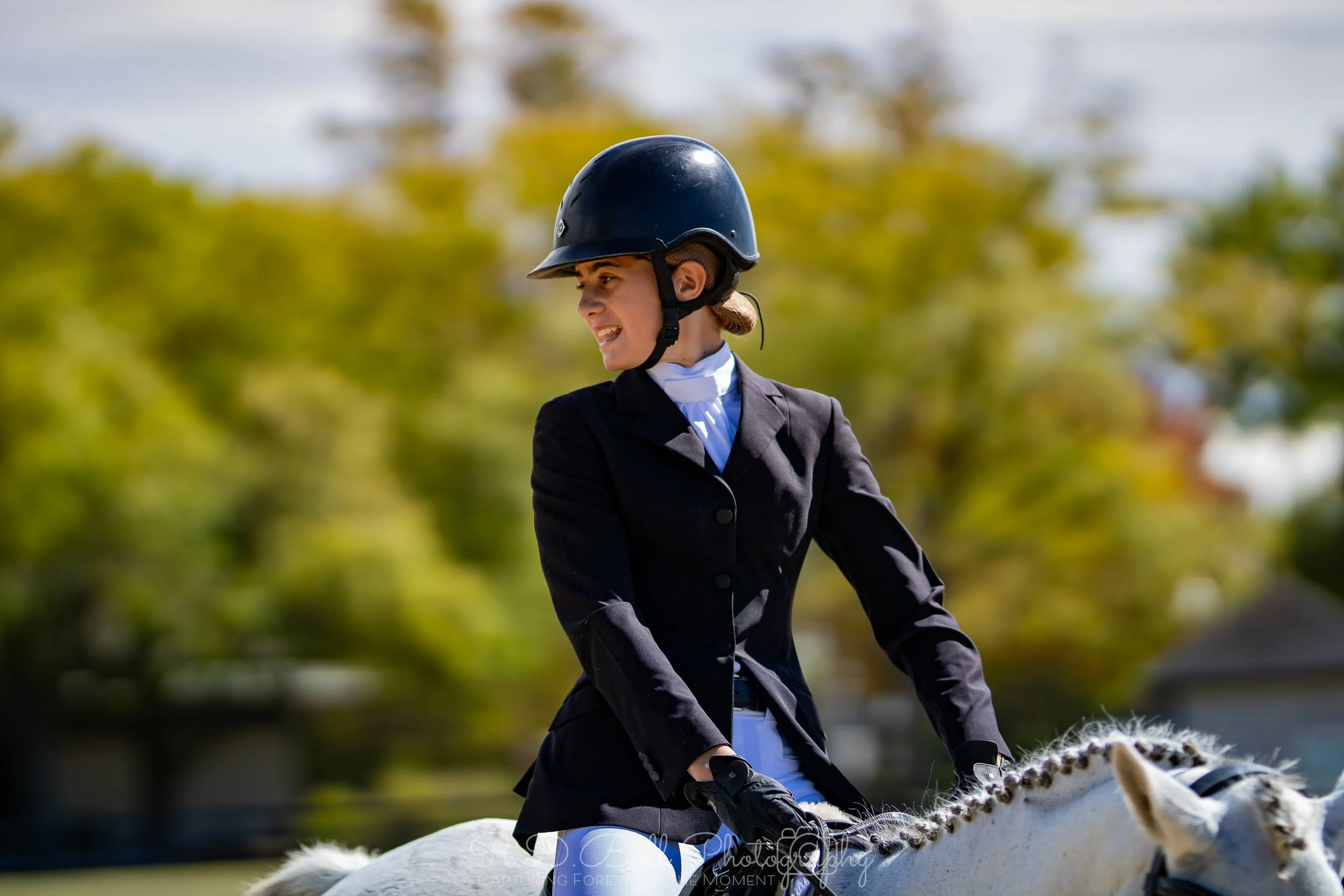 A woman in equestrian riding attire, including a black helmet and black riding jacket, riding a white horse outdoors with a blurred background of trees.