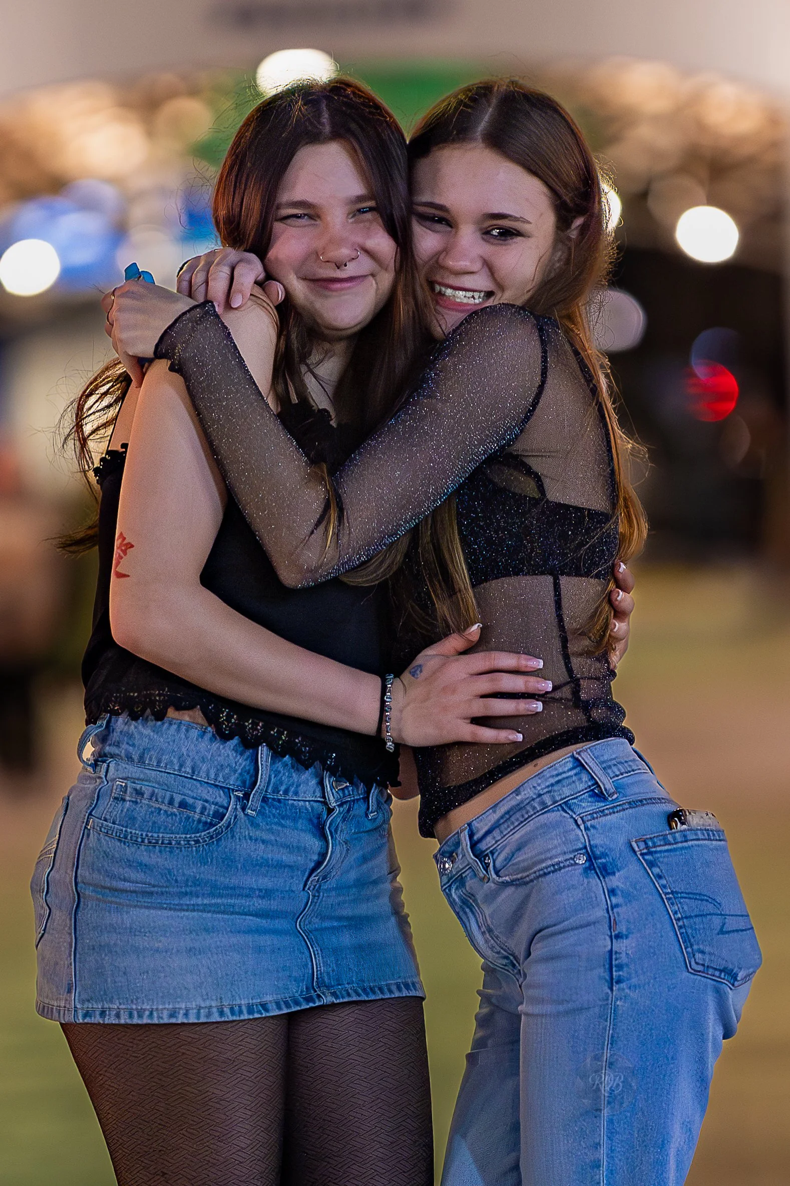 Two young women hugging and smiling at night, with a blurred background of city lights.