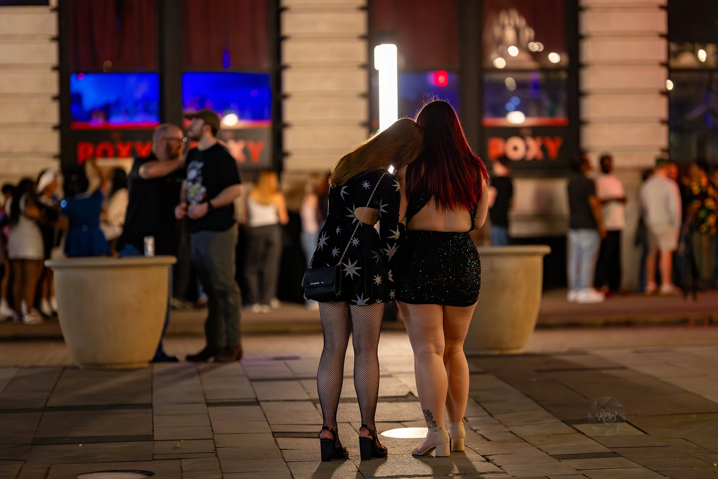 Two women with back to camera, one with red hair and sparkly dress, the other with blonde hair and black dress with star patterns, standing close and leaning towards each other outside at night with a crowd and a building in the background.