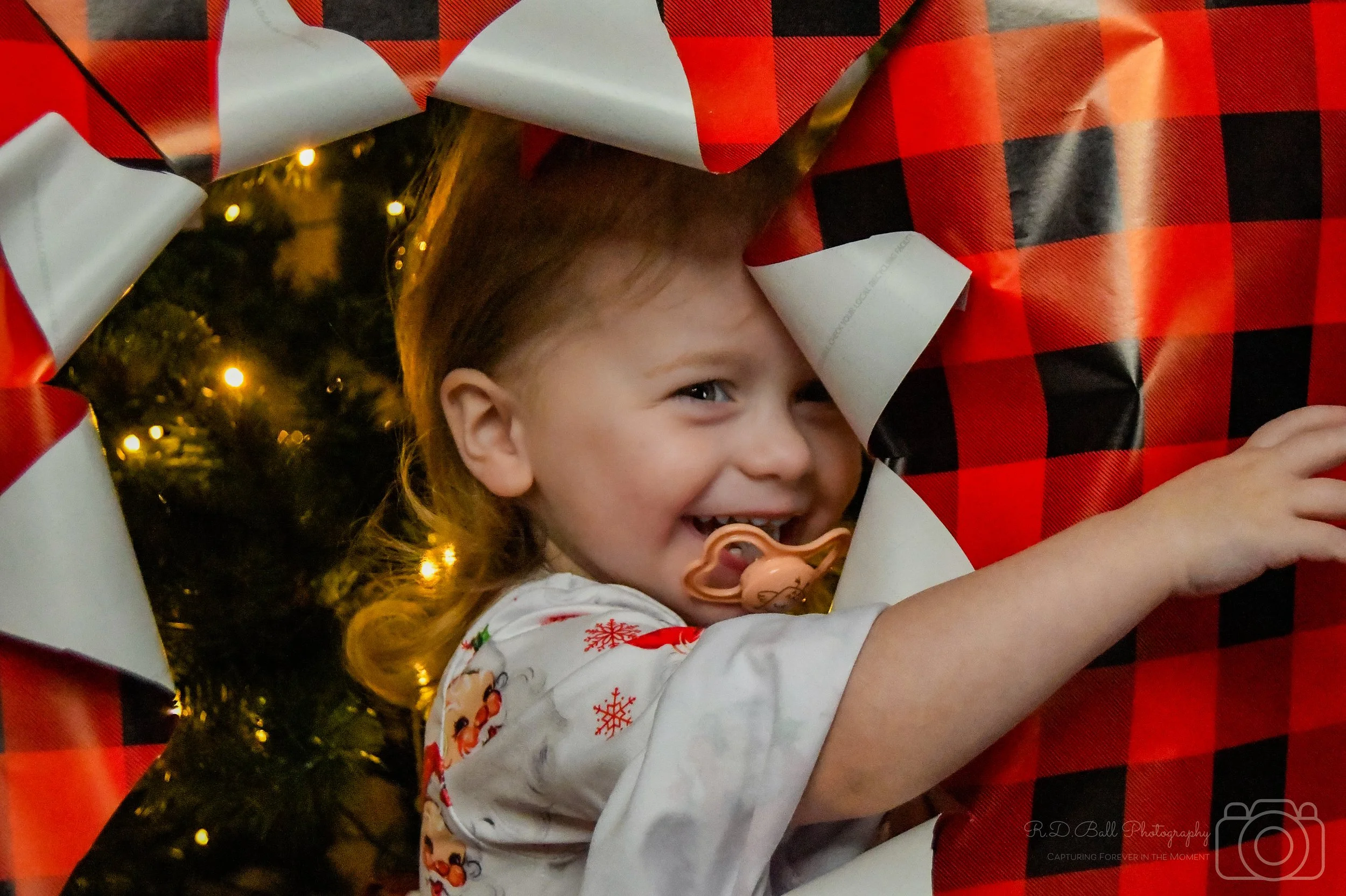 A young girl with light brown hair and a pacifier in her mouth, smiling as she peeks out from behind Christmas wrapping paper with red and black plaid pattern. In the background, there are Christmas tree lights.