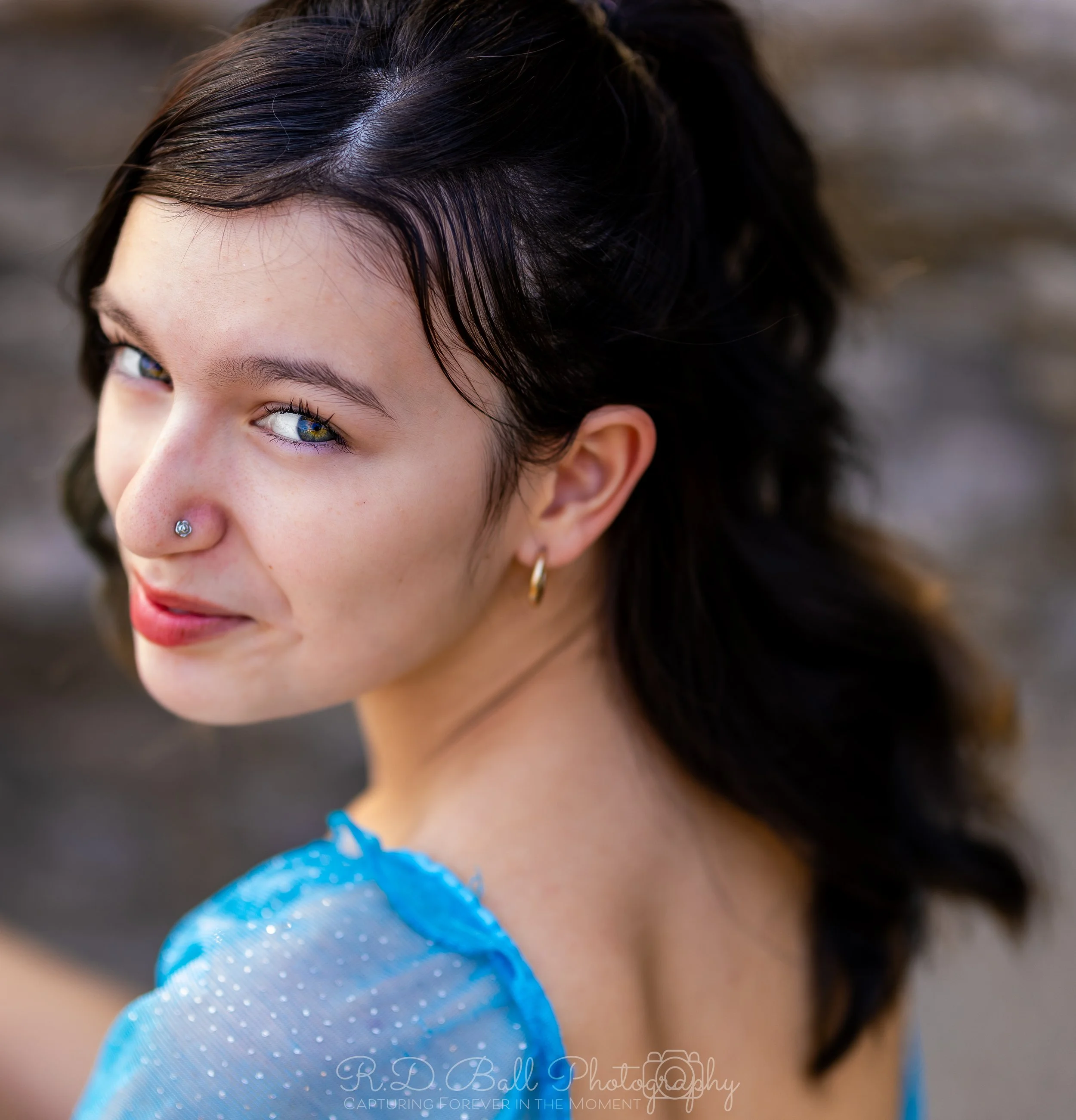 A young woman with dark hair, blue eyes, and light skin, looking over her shoulder with a slight smile, wearing a blue sheer top and gold hoop earrings, and a nose piercing.