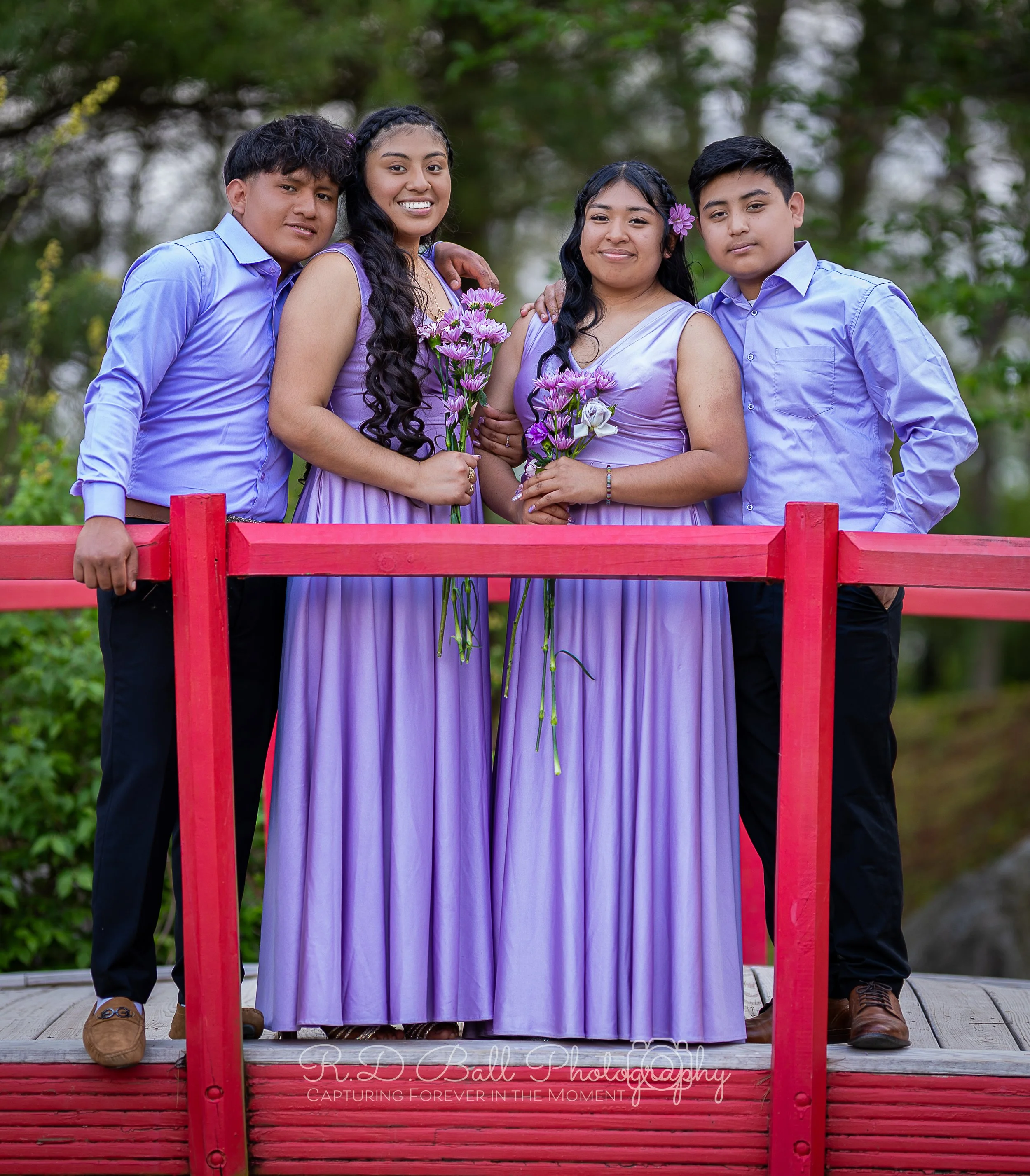 Group of five young people standing on a bridge with a red railing outdoors, wearing coordinated purple outfits, holding purple and white flowers, with trees in the background.