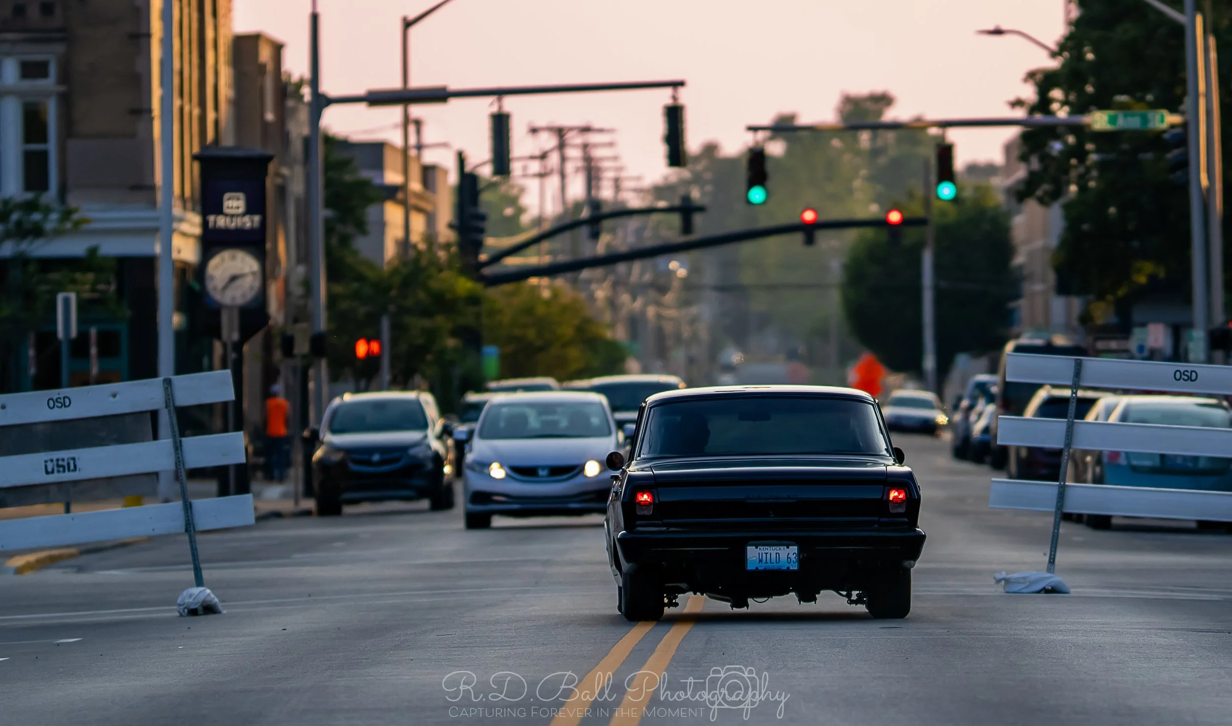 Black vintage car blocking a city street with traffic barriers, while other cars are stopped at traffic lights at dusk.