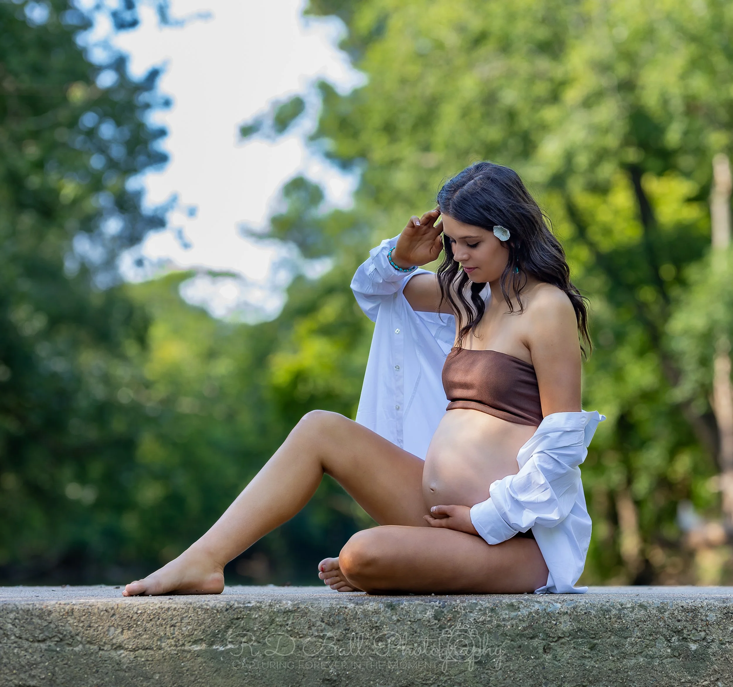 A pregnant woman sitting on a concrete surface outdoors, wearing a brown tube top and an open white shirt, with a woman in the background. The setting is lush and green, suggesting a park or forest.