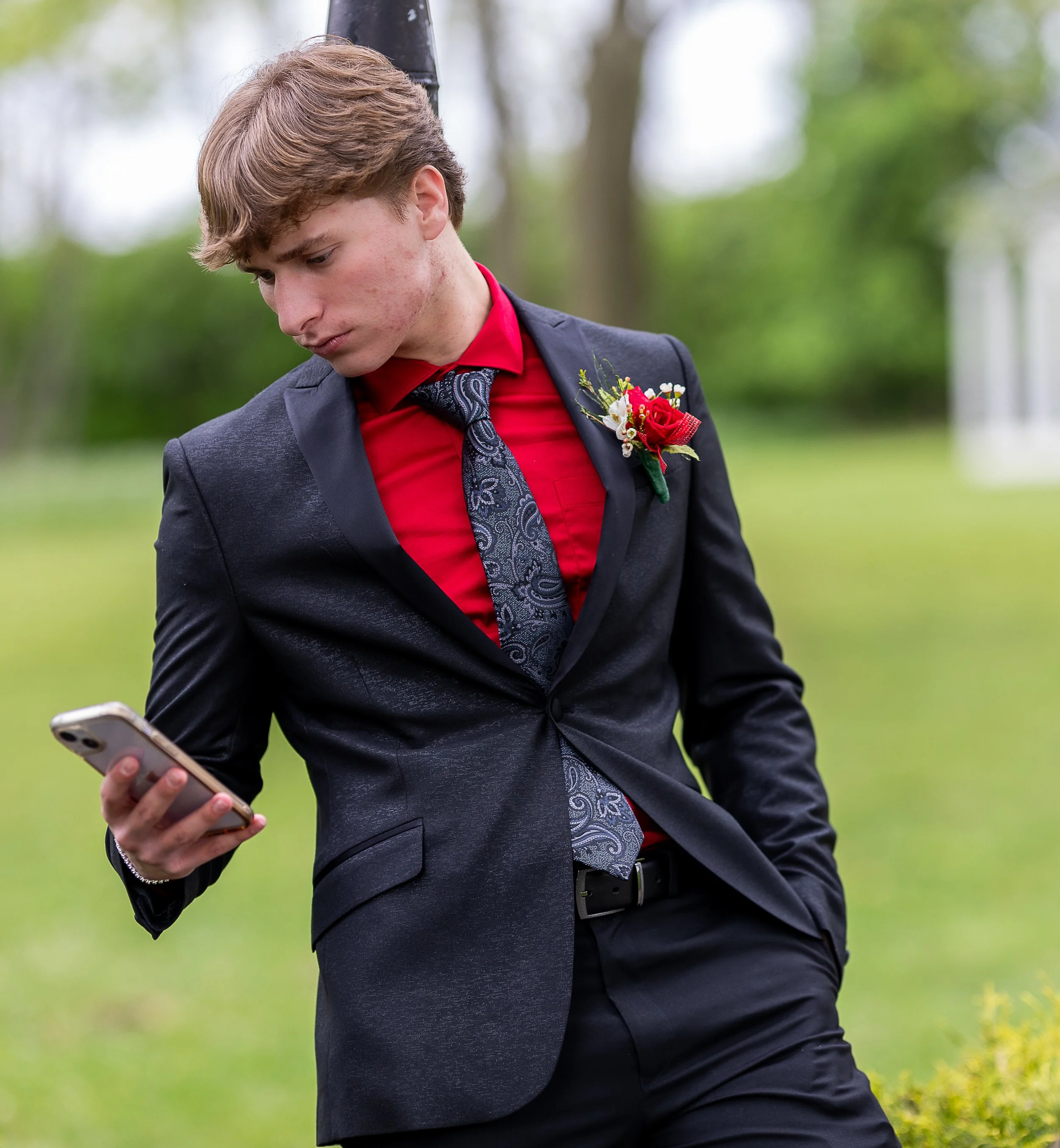 A young man in a black suit, red shirt, and patterned tie is looking at his phone outdoors in a grassy area with trees in the background.