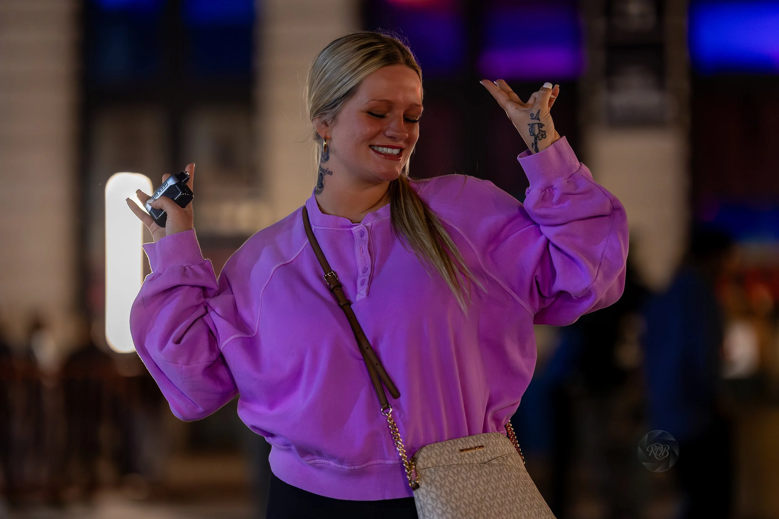 A woman with blonde hair wearing a purple sweatshirt dancing or celebrating indoors, holding a small black object in one hand, with a crossbody bag and tattoos visible on her neck and hands.