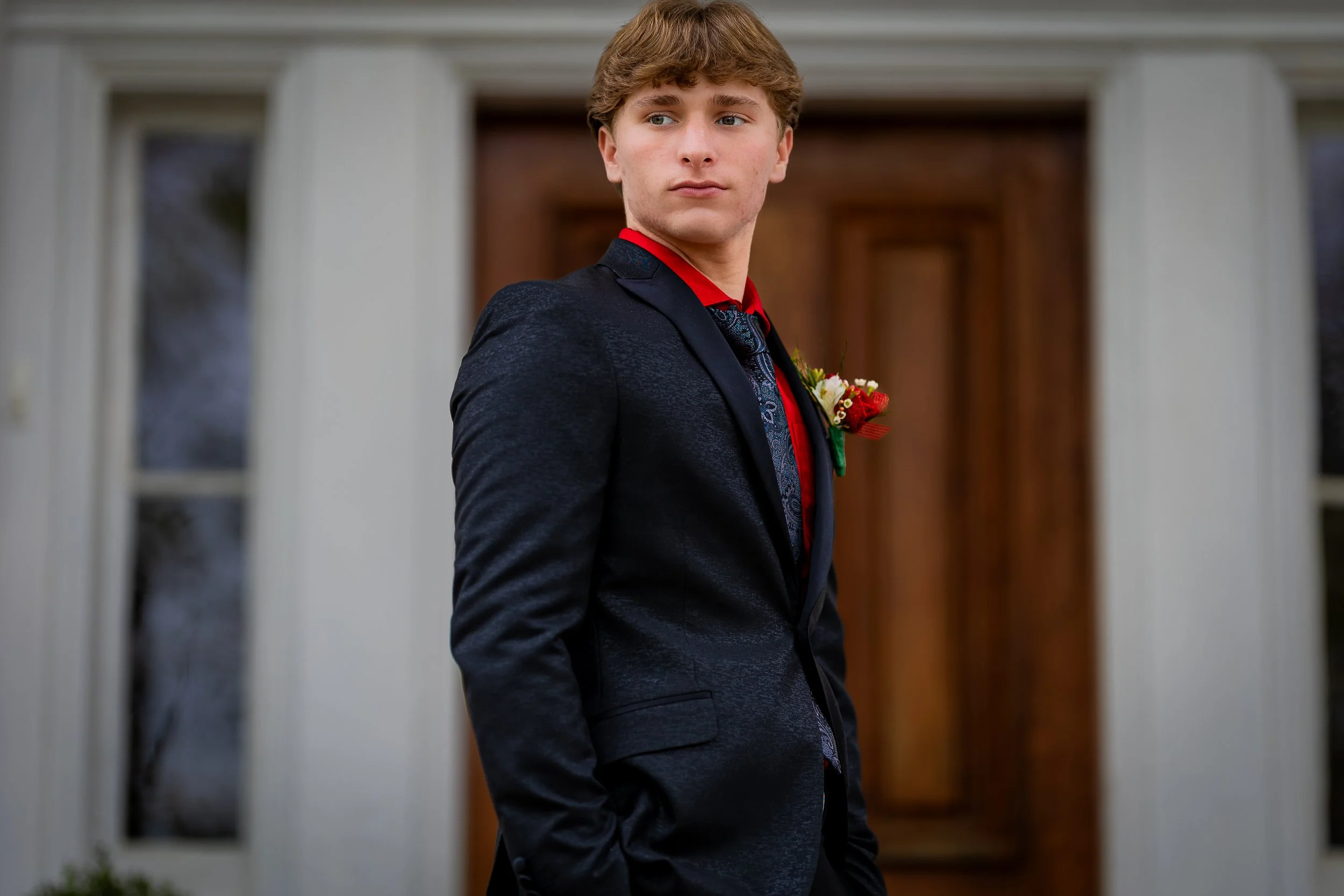 A young man in a dark suit with a red shirt and tie, standing in front of a house with a wooden front door. He has a boutonniere on his lapel.