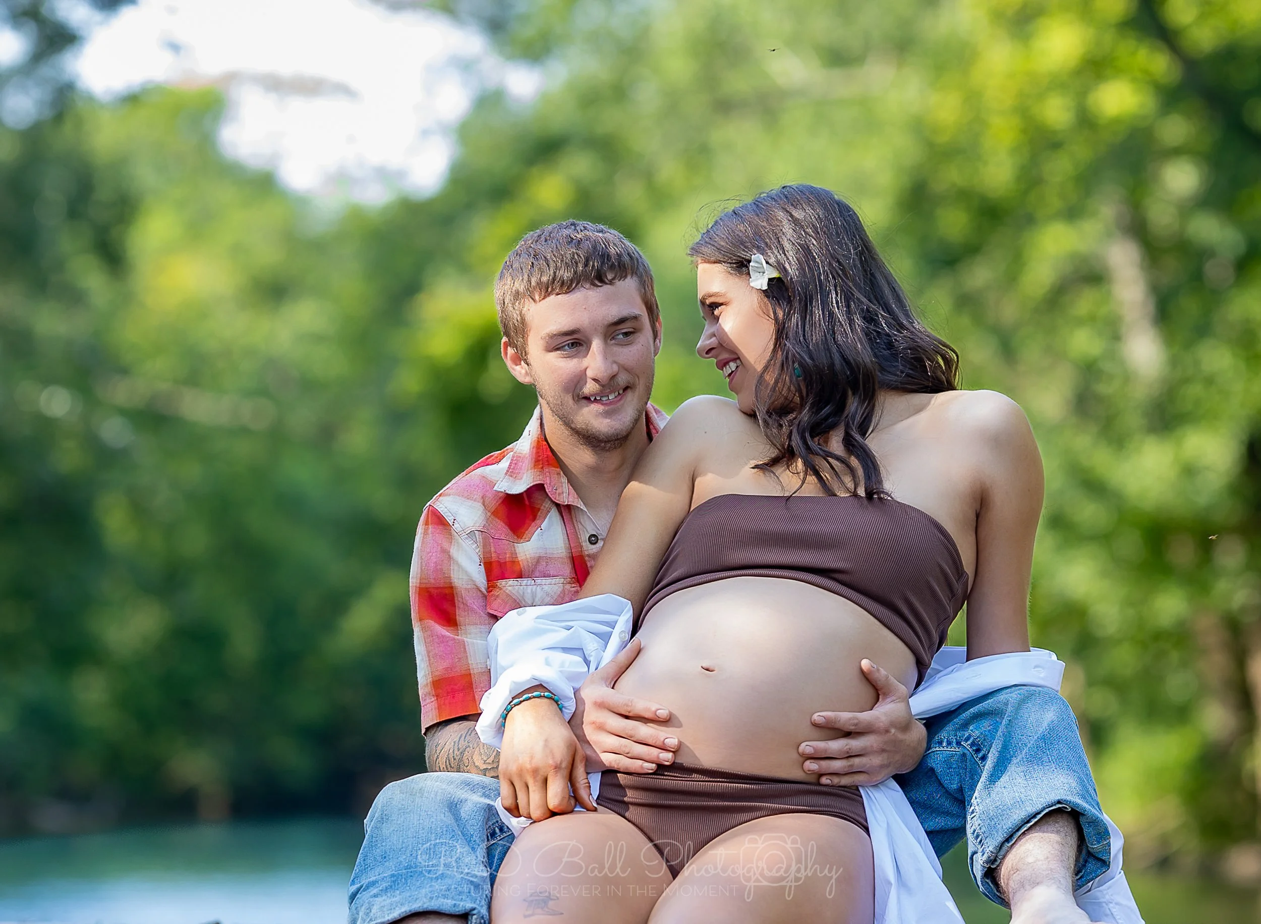 A happy couple outdoors with a woman who appears to be pregnant, smiling and touching her belly, while the man holds her and looks at her affectionately, surrounded by green trees.