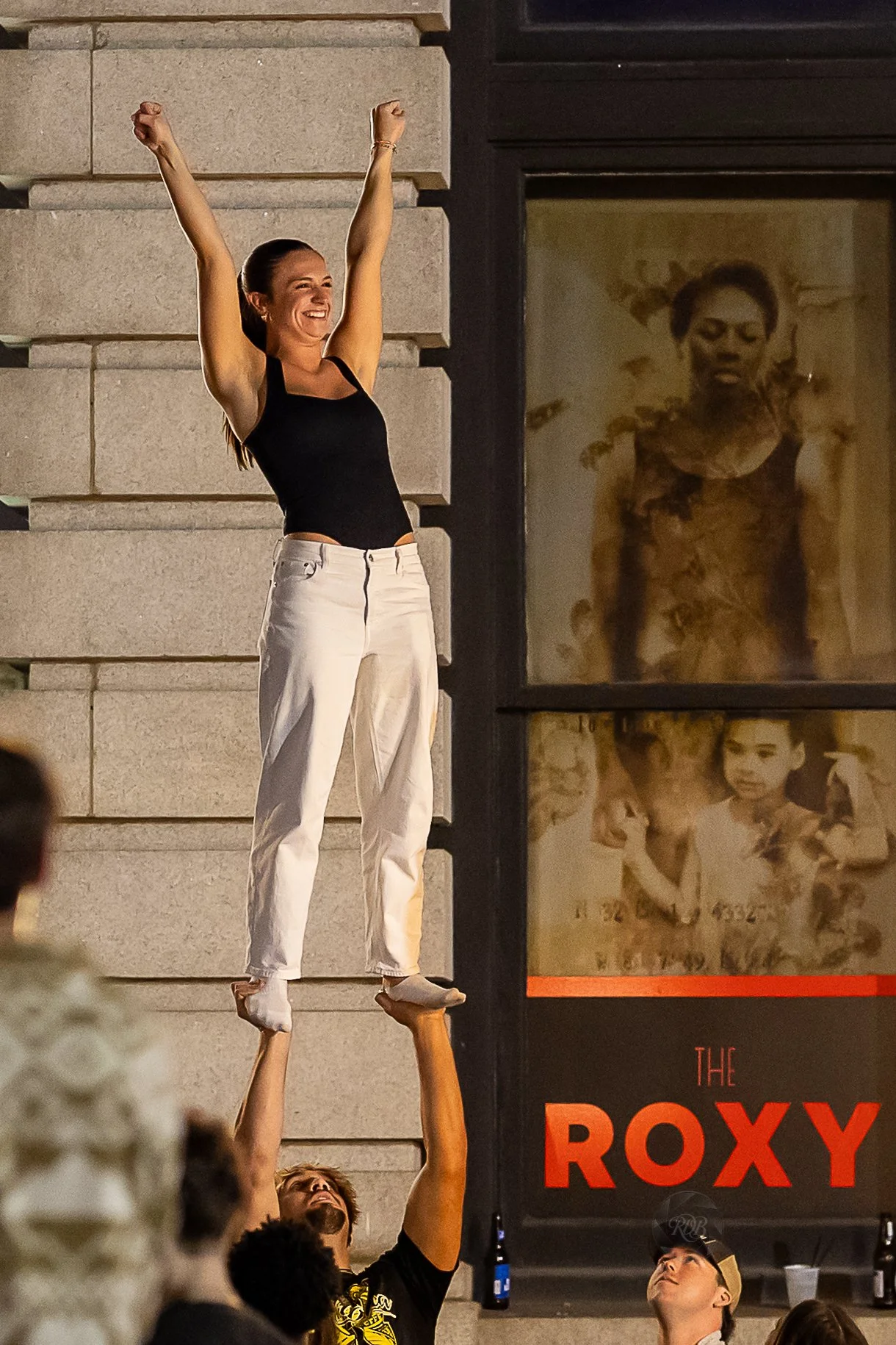 A woman in black top and white pants standing on the hands of a person, cheerfully celebrating, outdoors at night, with a storefront sign reading "The Roxy" in the background.
