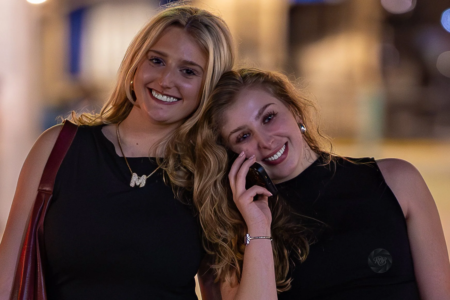 Two young women with blonde hair, wearing black dresses, smiling in an outdoor setting at night. One is on the phone, and they are standing close together.