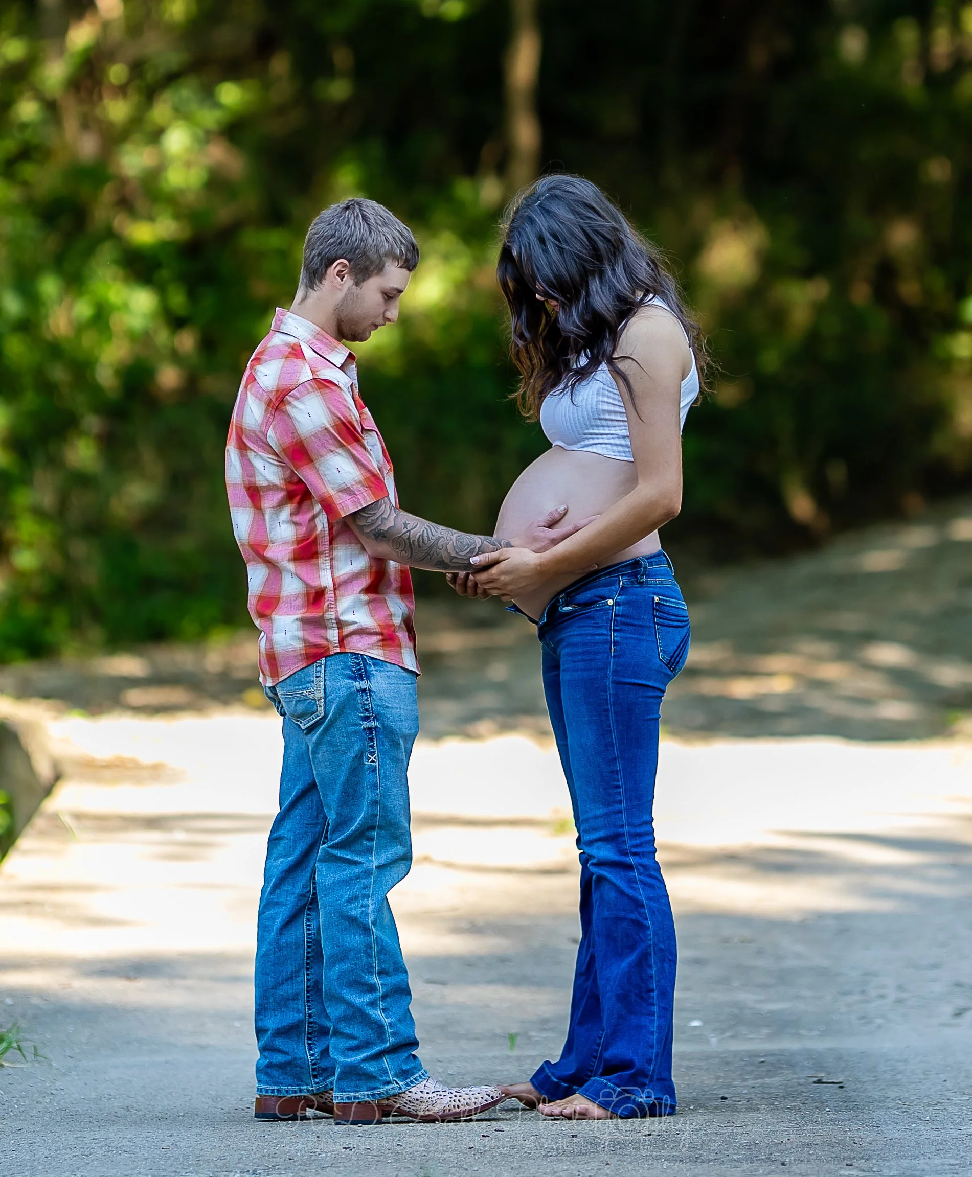 A pregnant woman with long dark hair in a white tank top and blue jeans stands barefoot outdoors on a dirt path, holding her belly. A man with short hair, wearing a red plaid shirt and blue jeans, gently holds her belly with both hands, looking down 