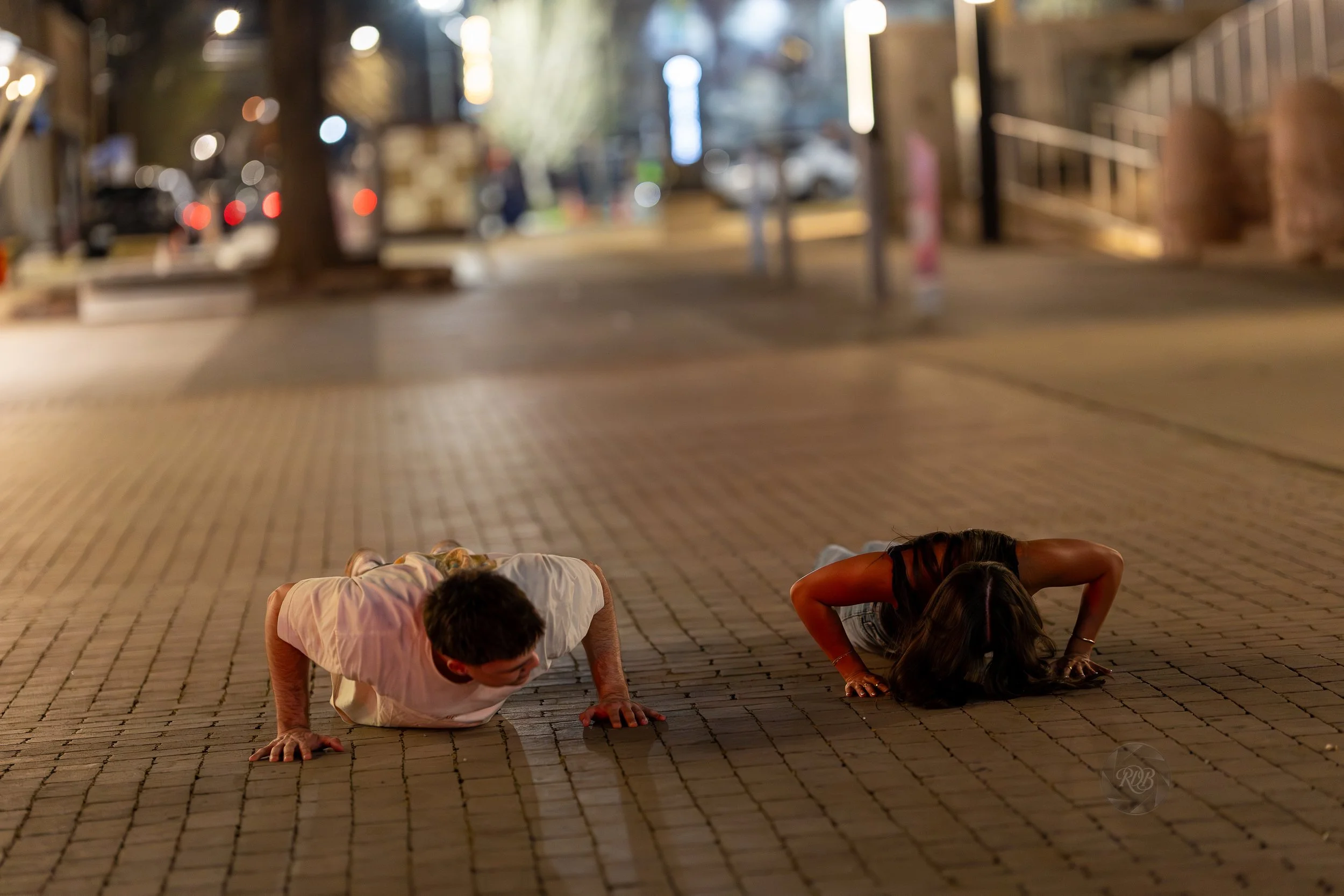 A man and woman performing push-ups on a brick sidewalk at night in an urban area.