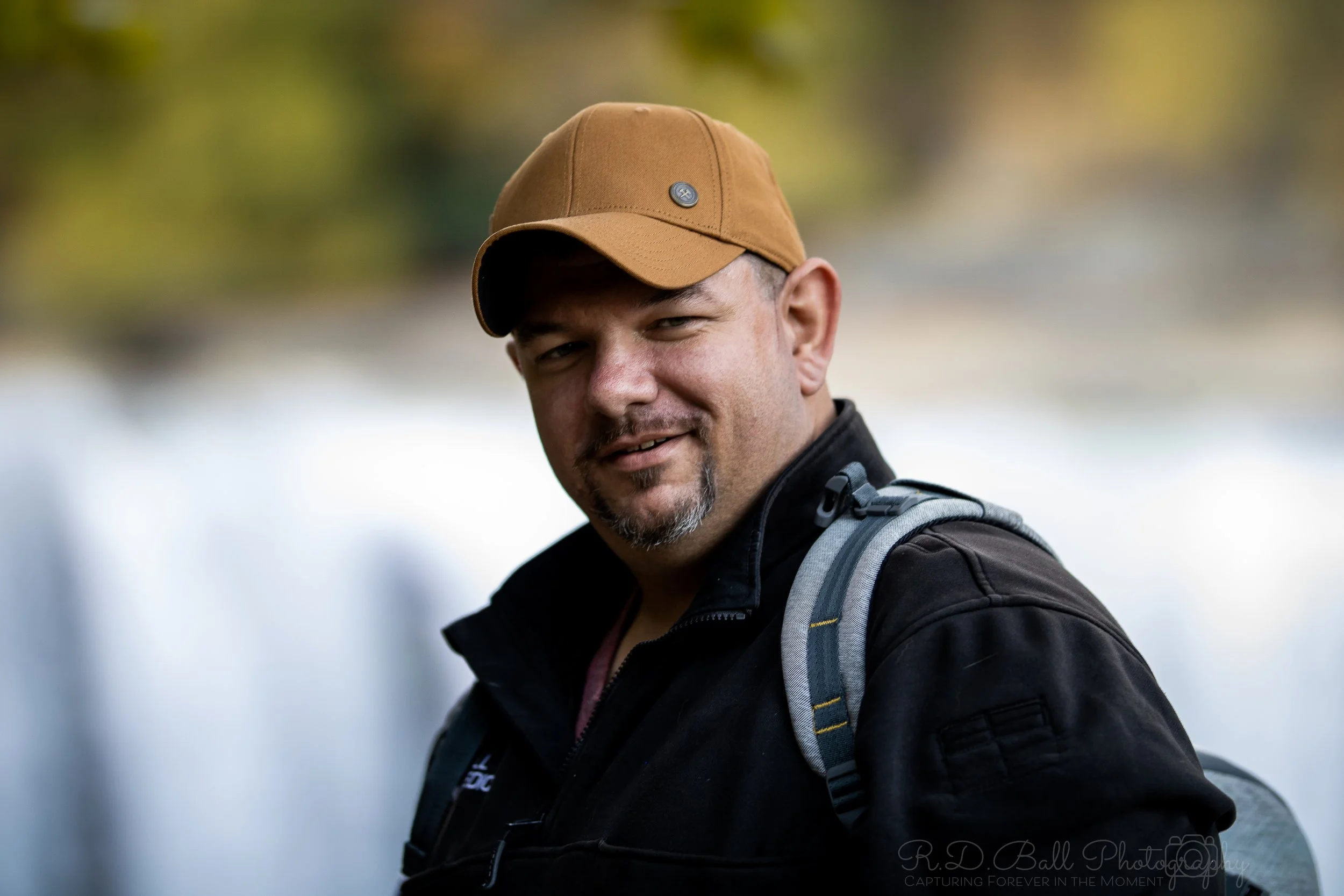 A man wearing a brown baseball cap, a black jacket, and a gray backpack outdoors, with a blurred natural background.