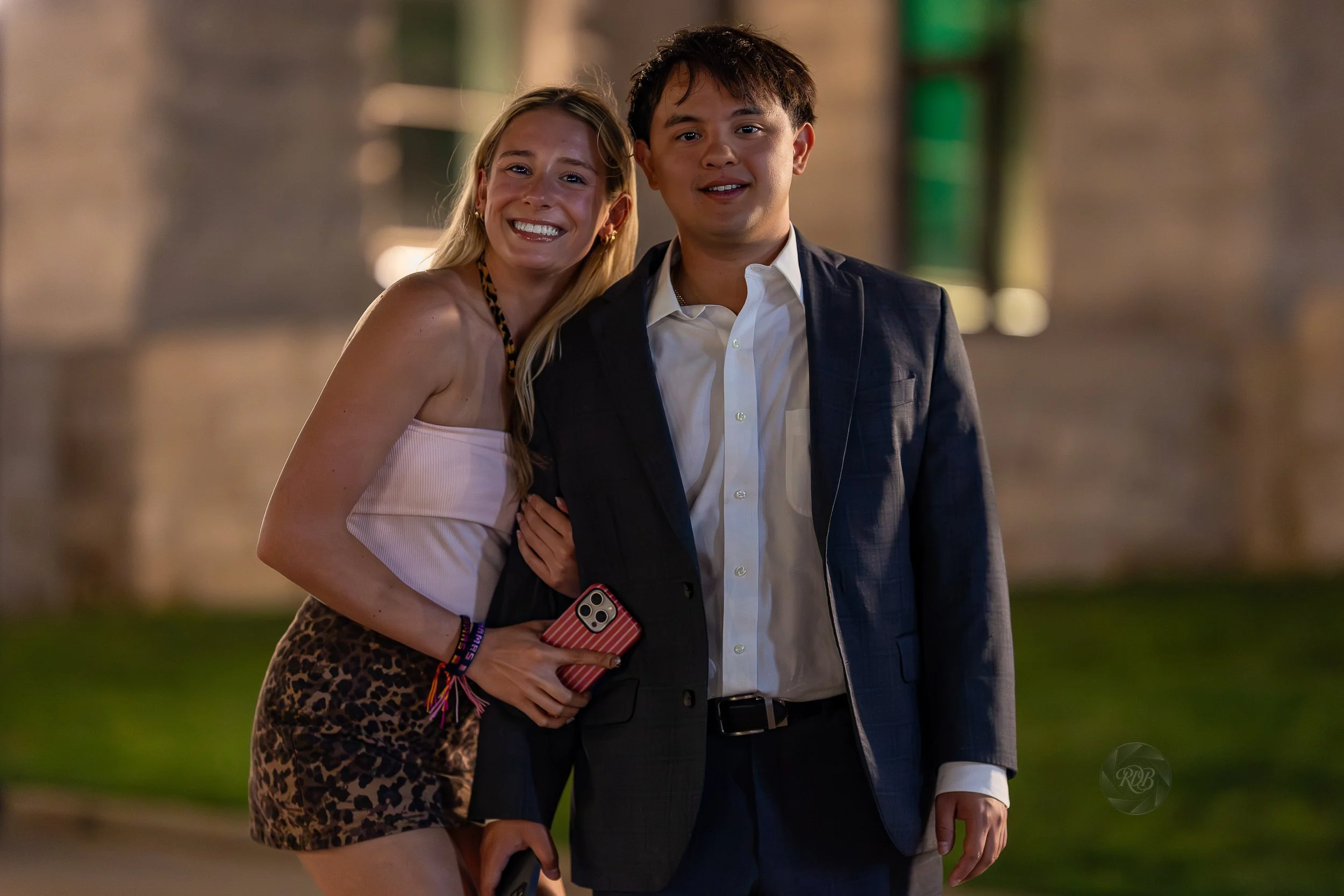 A woman in a white top and leopard print skirt and a man in a suit are standing outdoors near a brick building at night, smiling for the camera.