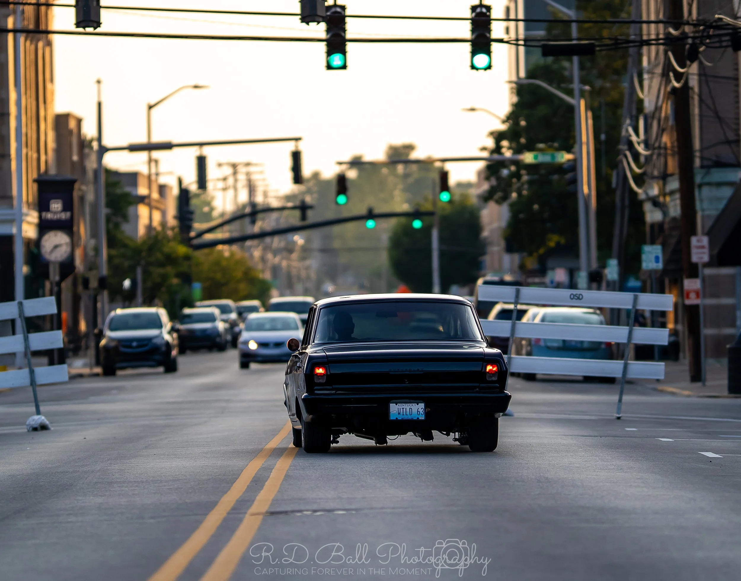 Black vintage car stopped in the middle of a city street with green traffic lights overhead and several cars in the background.