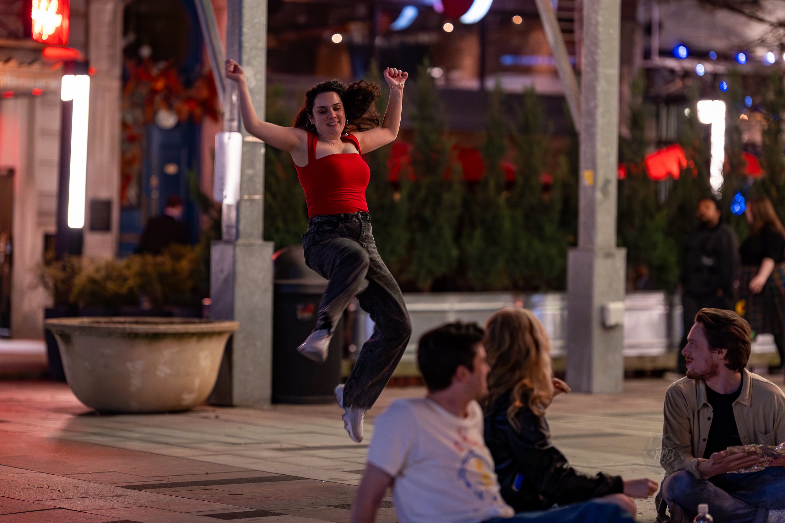 A woman in a red tank top and black jeans jumping in the air with arms raised in a lively outdoor area at night. Four people are sitting on the ground nearby, and there are colorful lights and trees in the background.