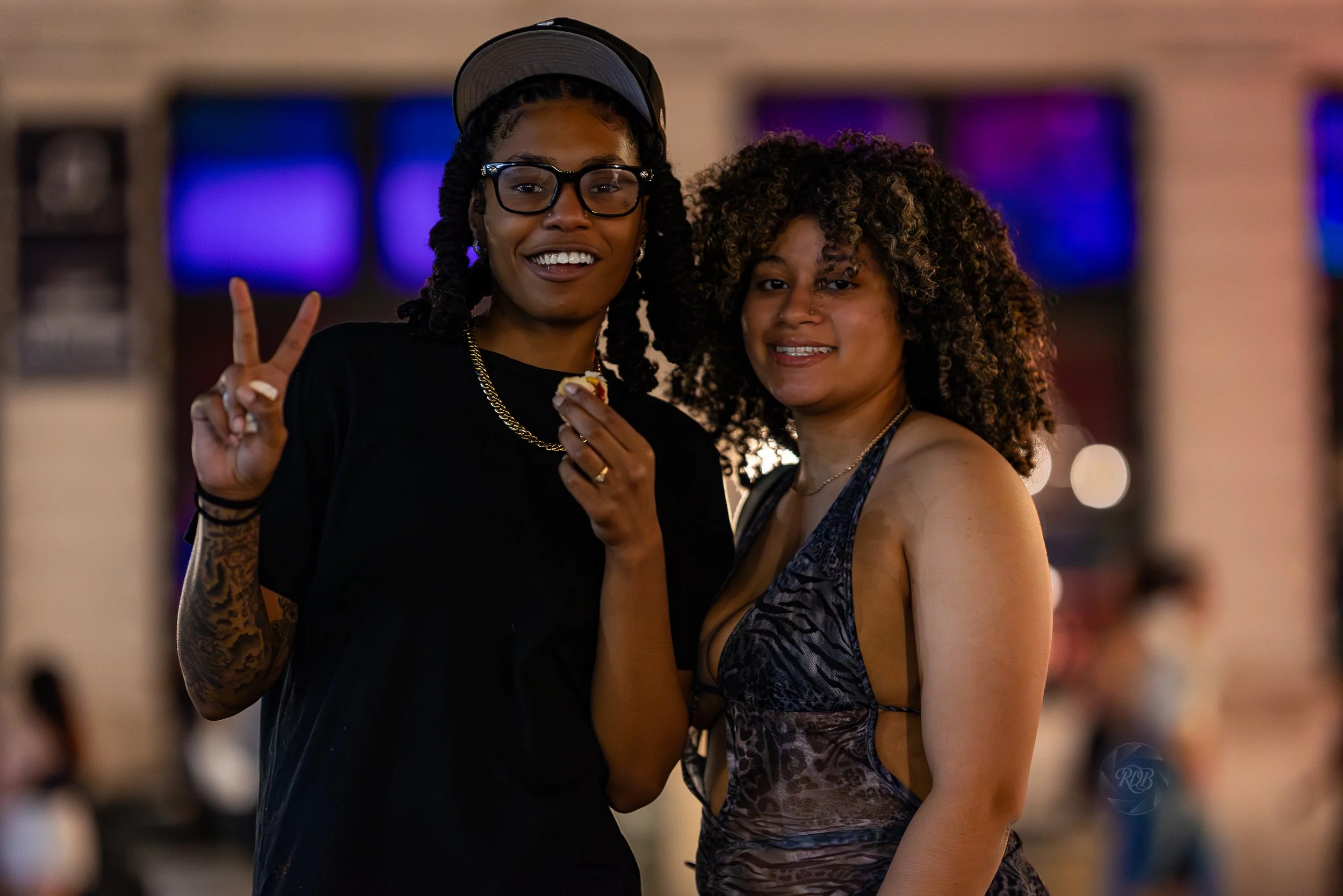 Two women smiling at the camera in a social setting, one making a peace sign with her fingers, the other holding a small food item.