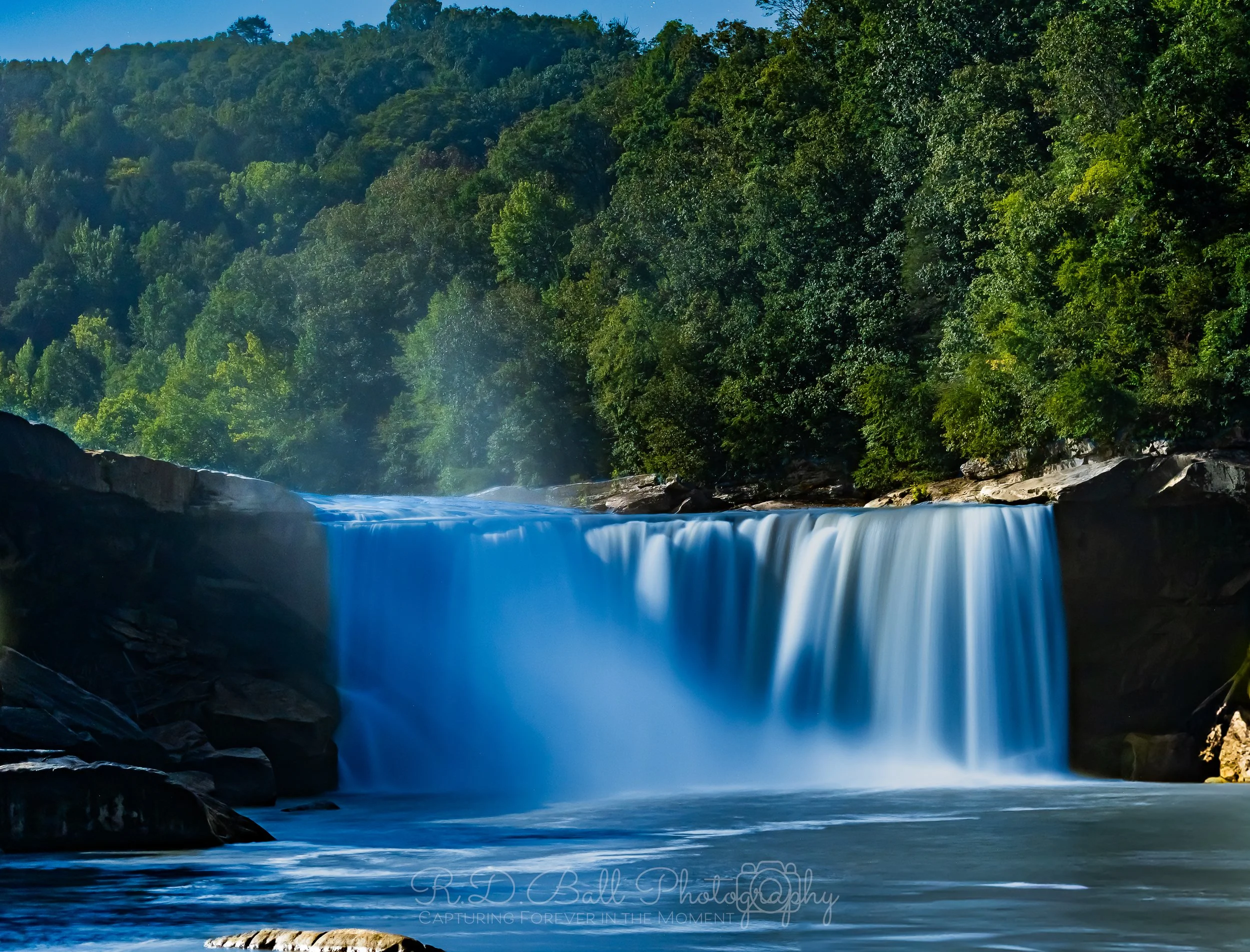 A river waterfall cascading over rocks surrounded by lush green trees in a forested area.