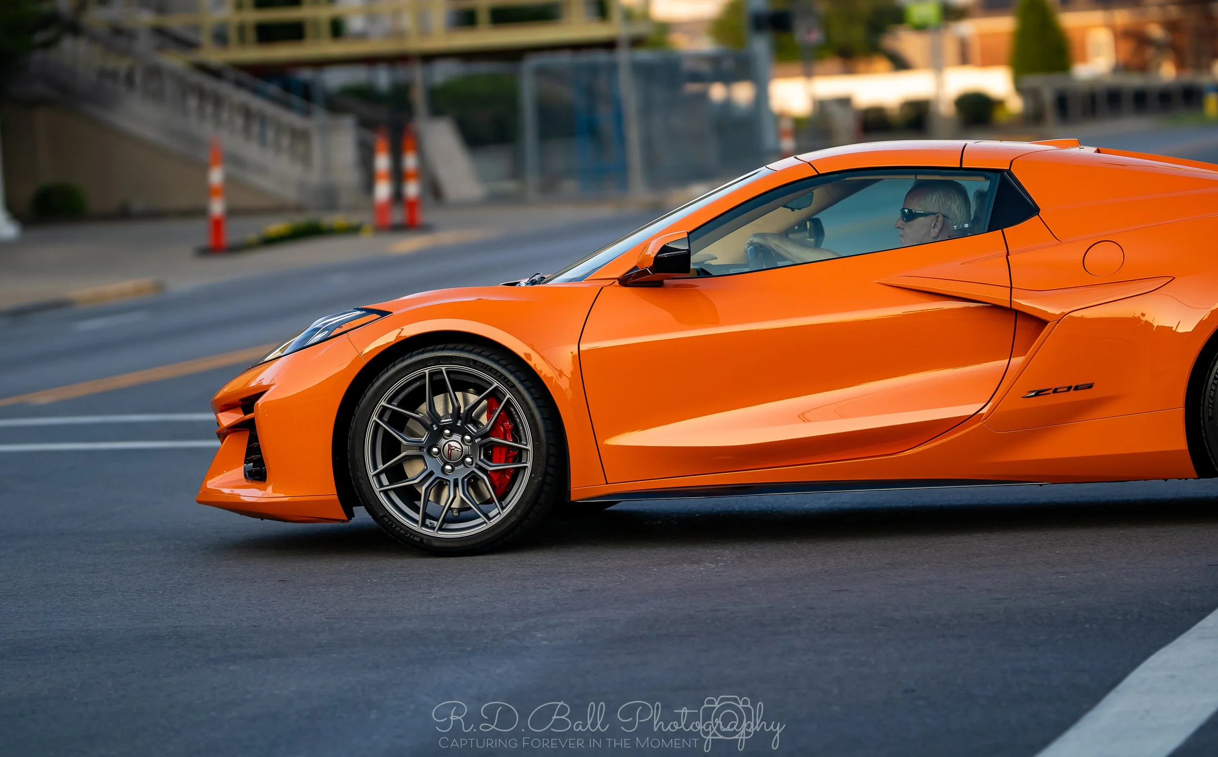 An orange sports car driving on a city street with a driver wearing sunglasses inside.