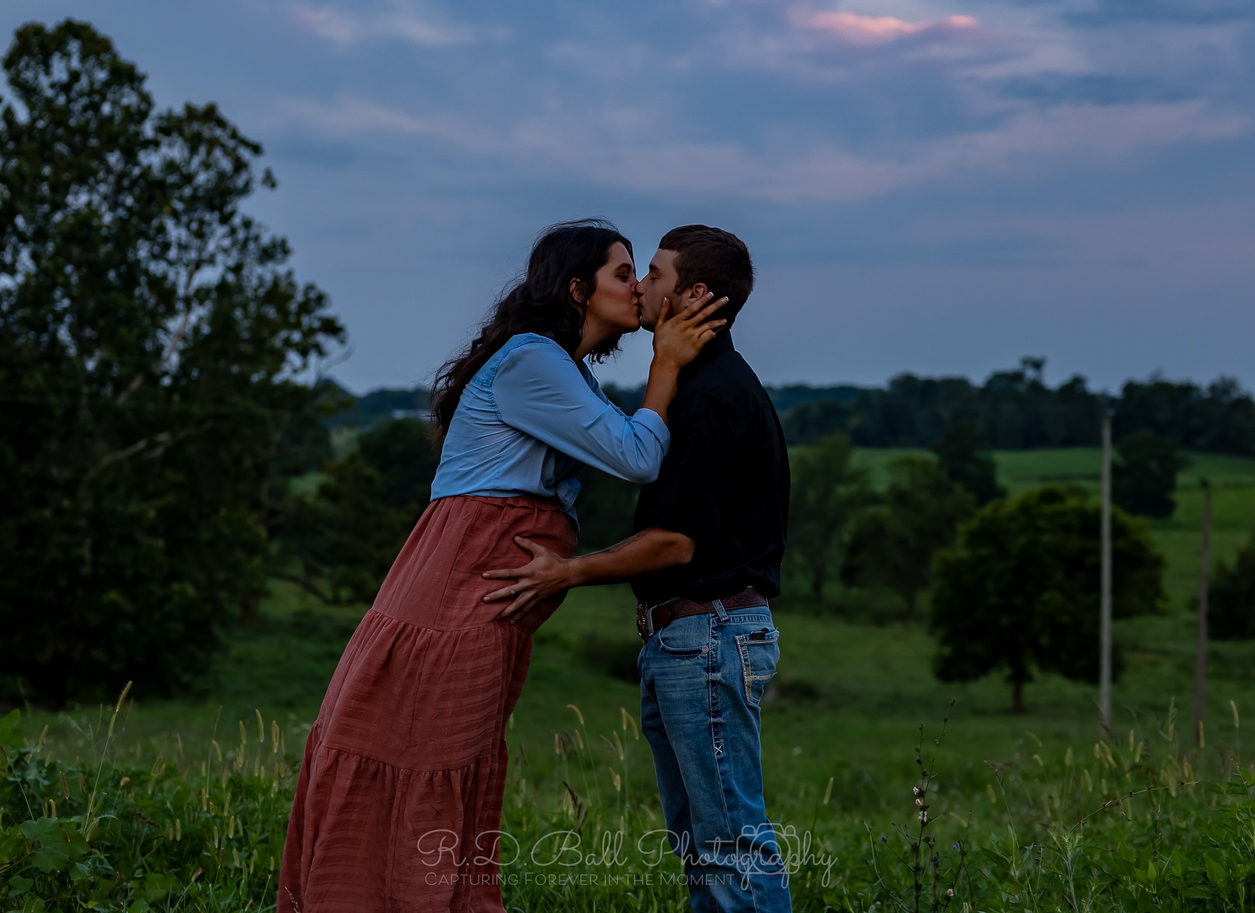 A couple kissing outdoors on a grassy field with trees and hills in the background, cloudy sky above.
