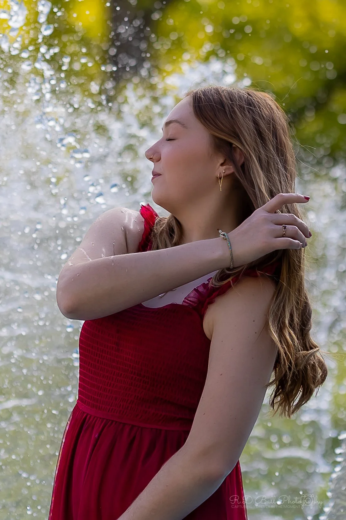 A woman in a red dress with wet hair stands outdoors with water droplets on her arm, eyes closed, and a peaceful expression.