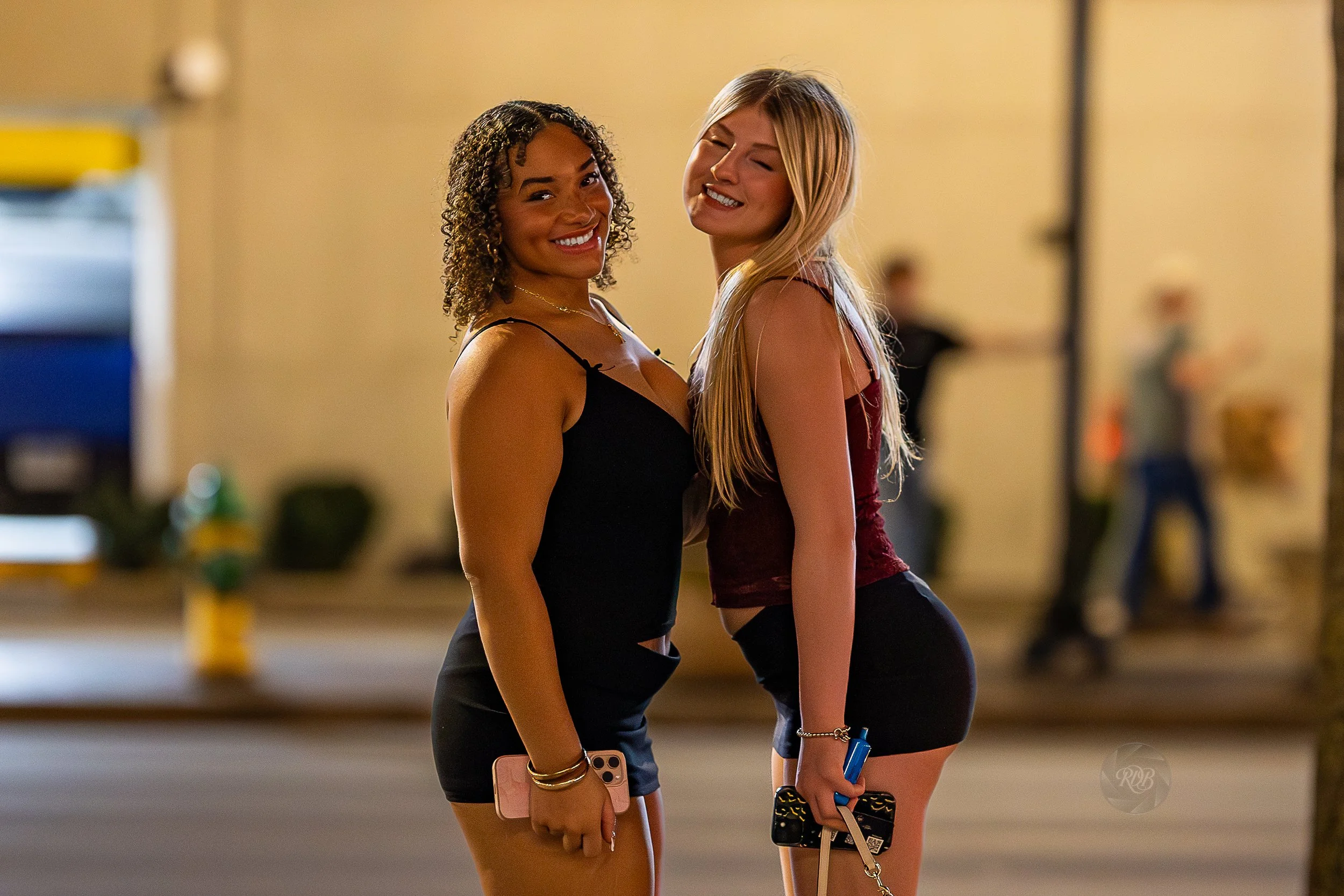 Two young women with smiles standing close together on a city street at night, both holding smartphones and dressed in casual summer outfits.