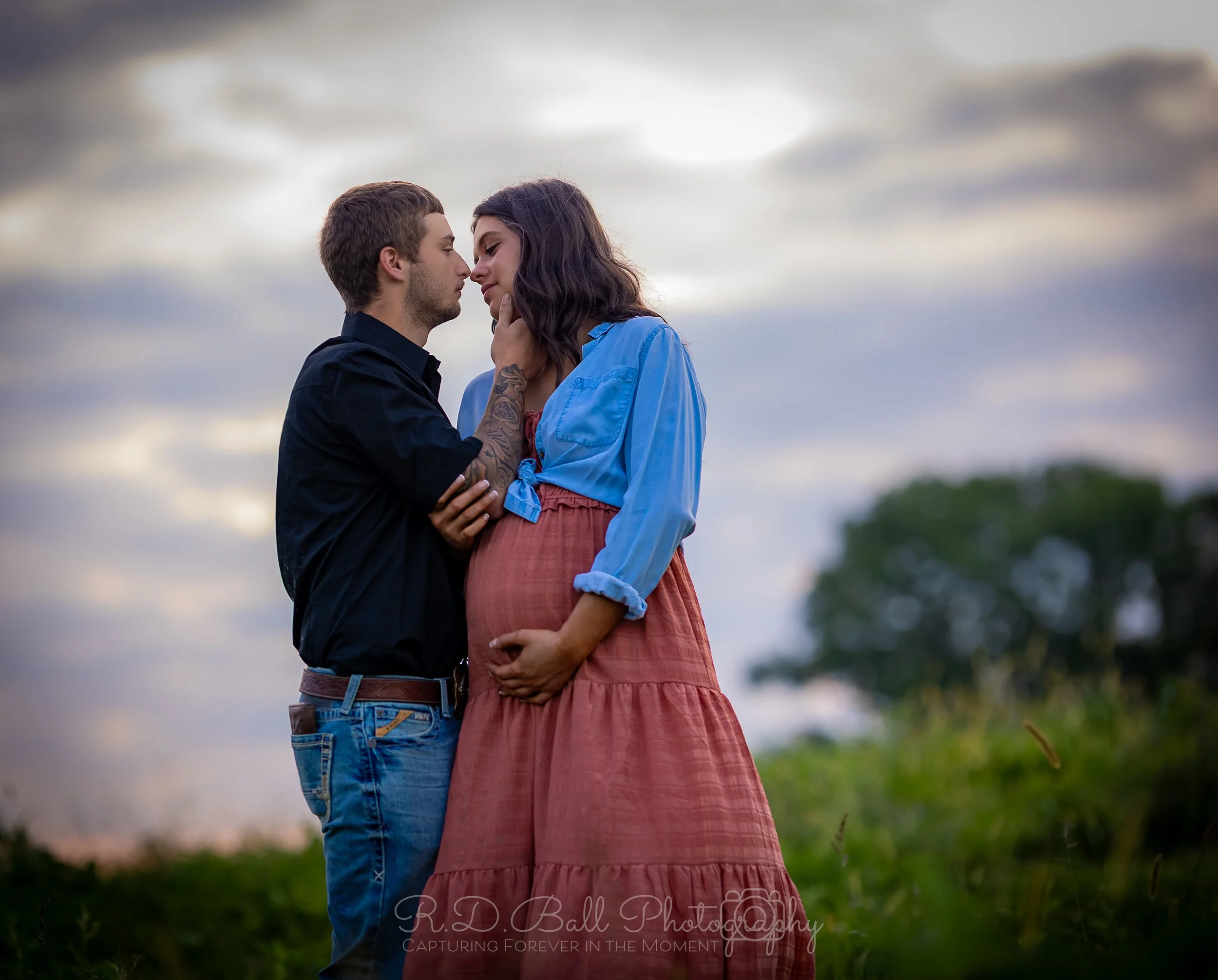 A pregnant woman with long brown hair and a man with short brown hair standing close together outdoors, touching foreheads and holding each other's faces, with a cloudy sky and trees in the background.