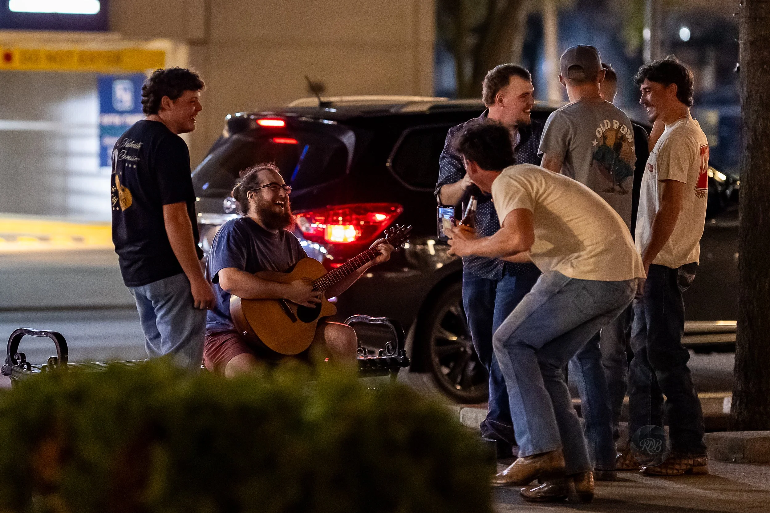 Group of people singing and playing guitar on a sidewalk at night, with a black car and a parking lot in the background.