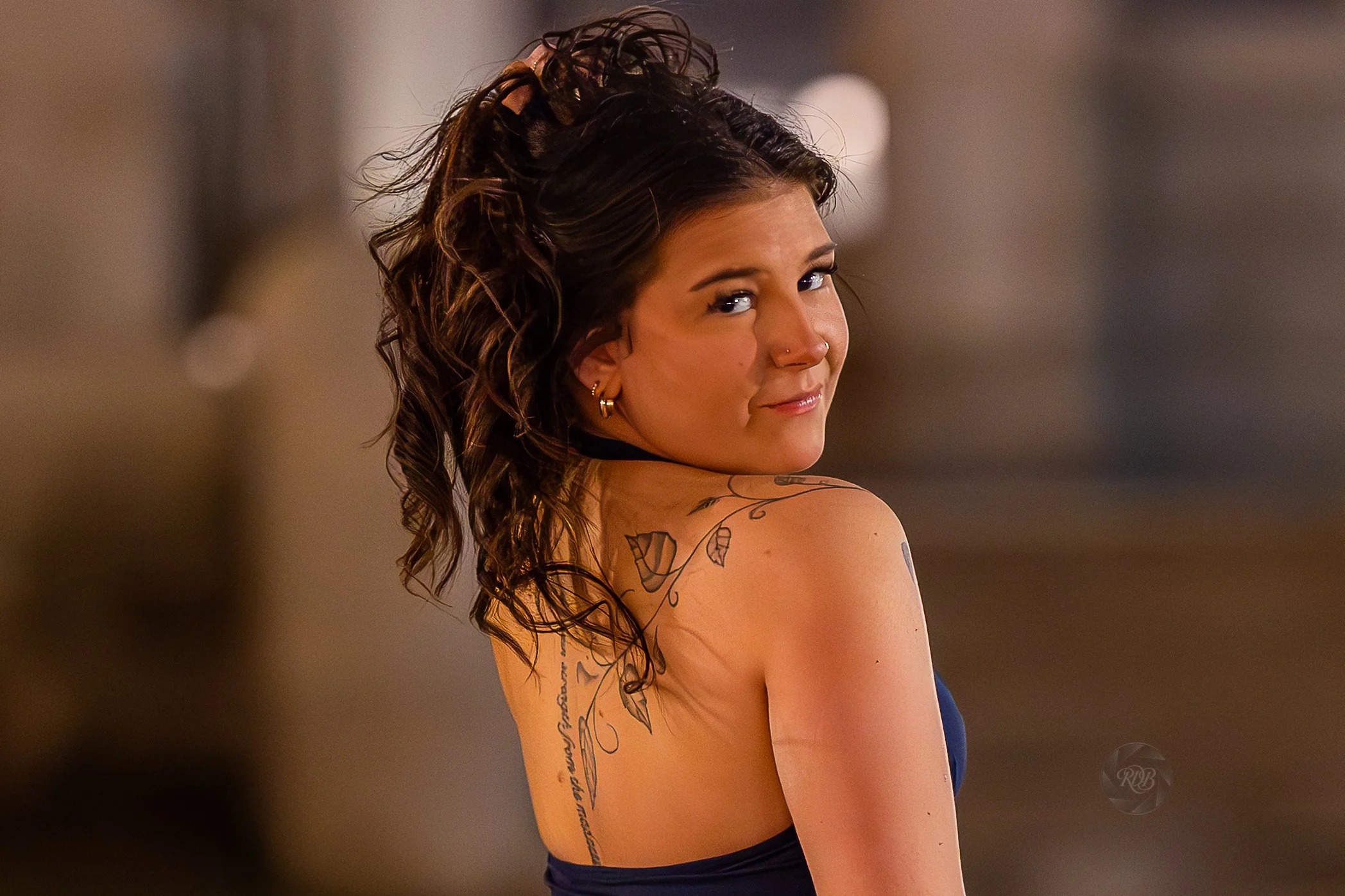 Young woman with wavy dark hair, tattoos on her shoulder and back, wearing a strapless dress, looking over her shoulder with a smirk.