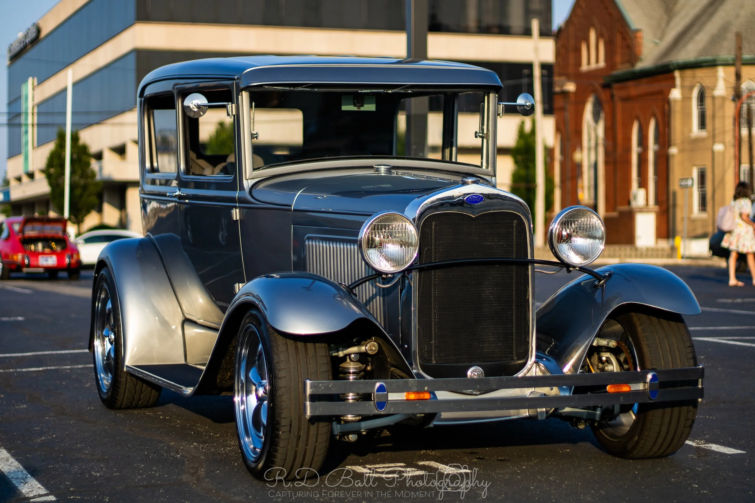 A vintage black and silver Ford car parked in an outdoor lot with modern and old buildings in the background.