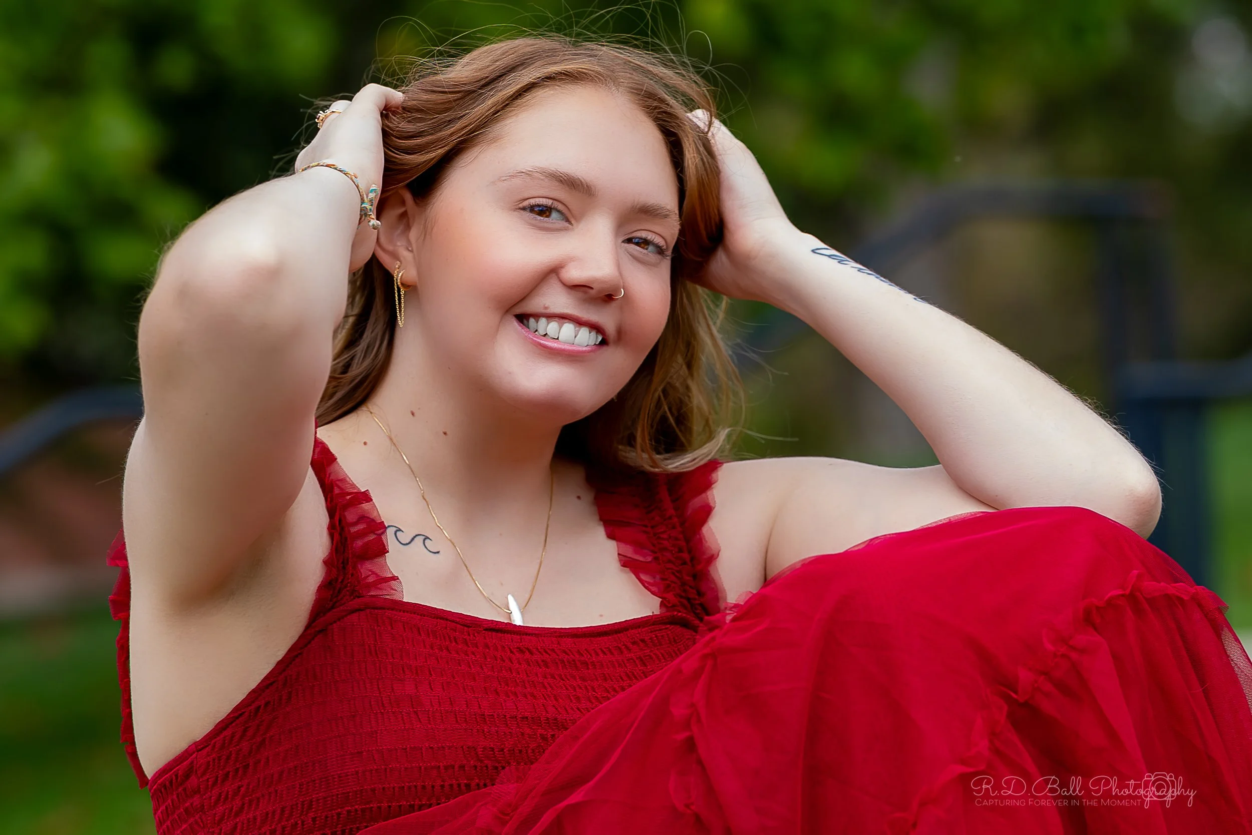 Smiling woman with light brown hair, wearing a red dress, jewelry, and a nose ring, outdoors with blurred green foliage in the background.
