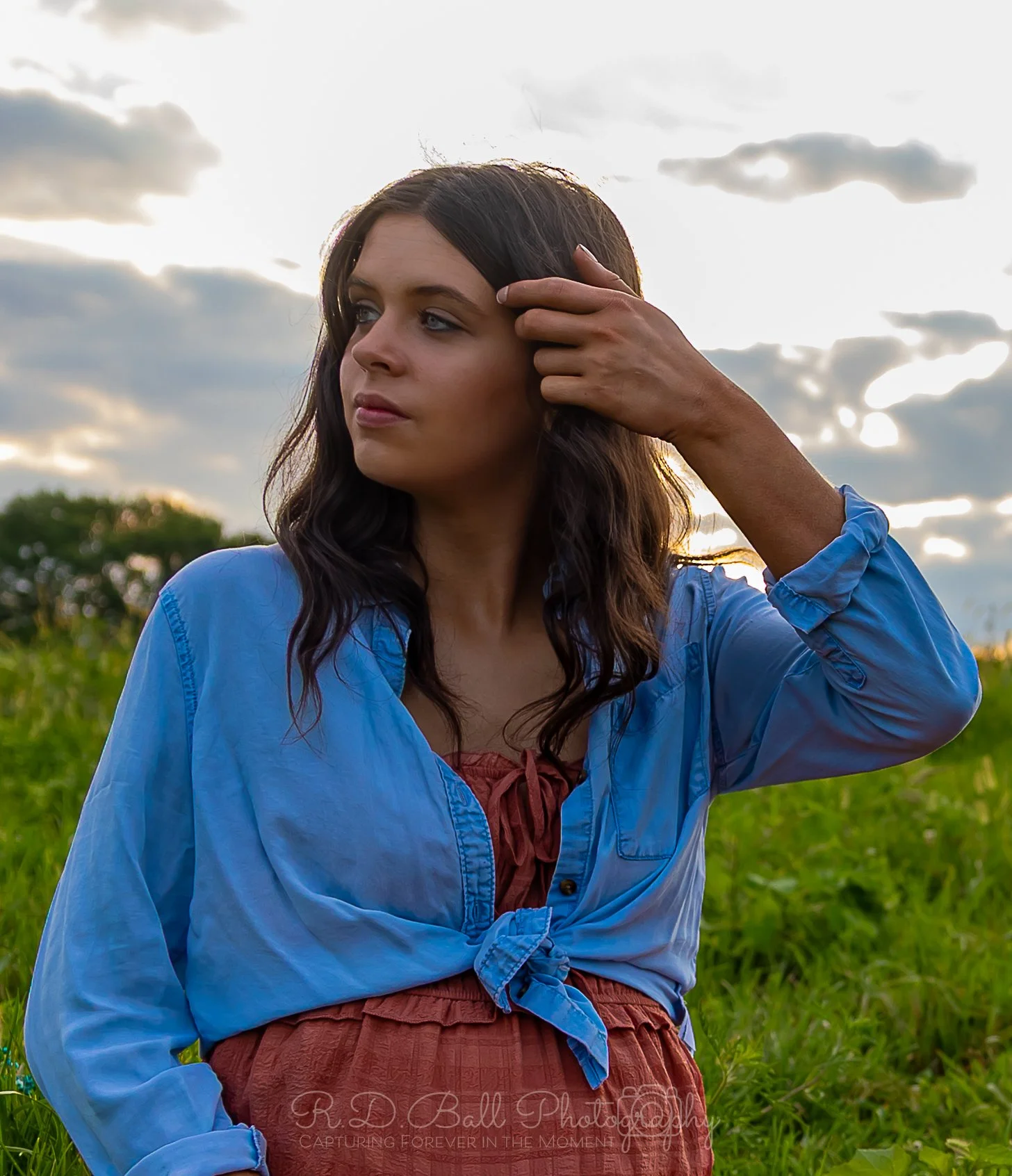 A woman with dark brown hair and blue eyes wearing a blue shirt and a reddish-brown dress outdoors during sunset, sitting in a grassy field with a cloudy sky in the background.