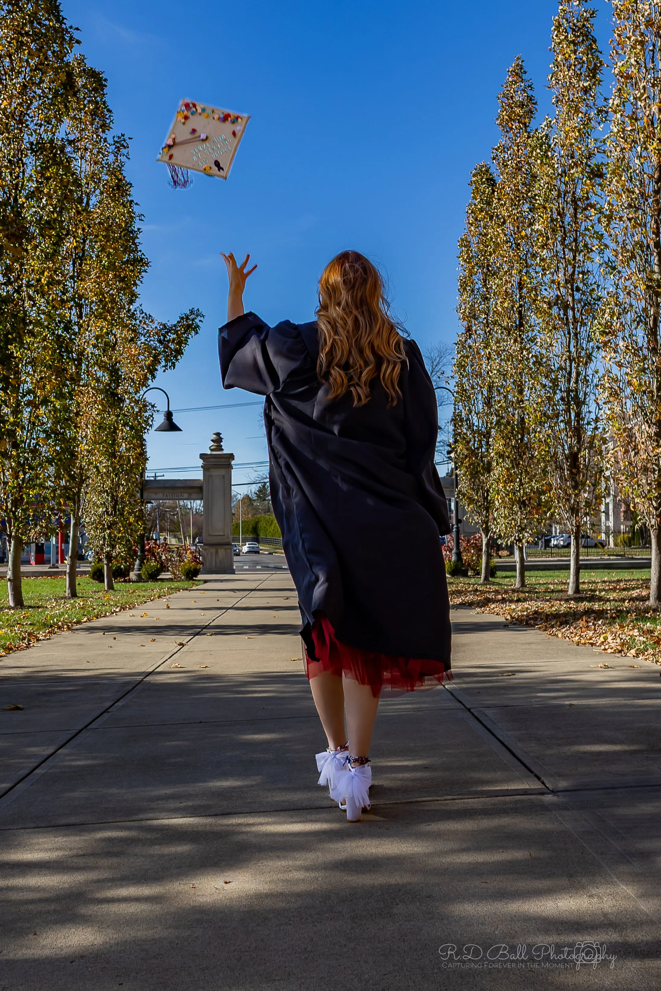 A woman in a graduation gown walking on a sidewalk outdoors, throwing her cap in the air.