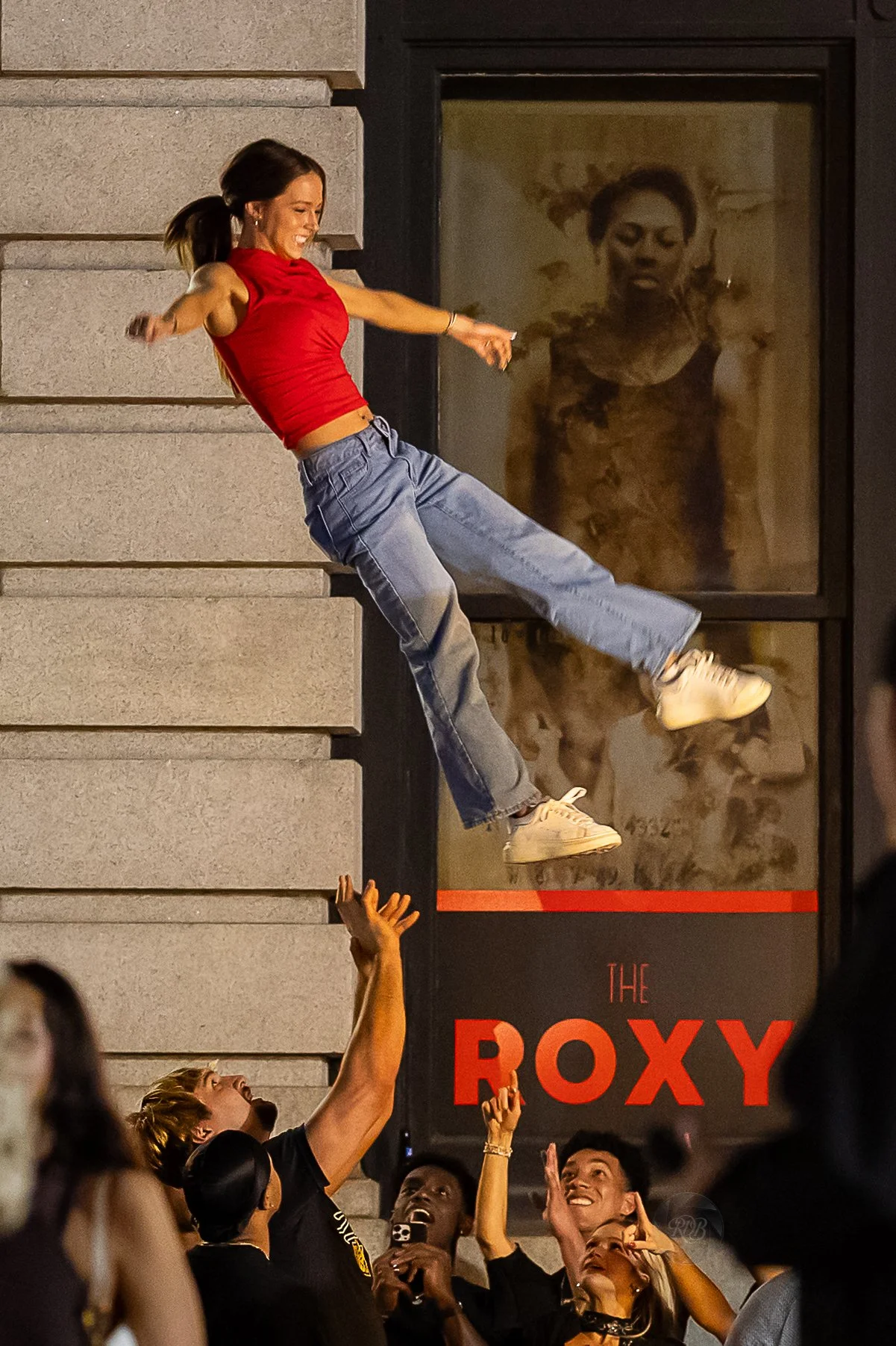 A young woman in a red tank top and blue jeans balances in mid-air leaning against a building's wall during a street event at night, with several people below reaching up or observing her. The scene appears lively and energetic, with a sign for The R