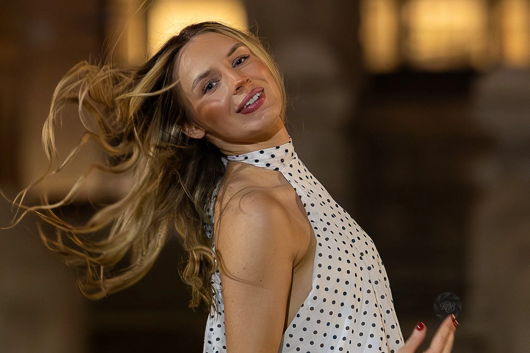 A woman with long wavy hair flipping her hair, wearing a sleeveless polka dot blouse, and smiling.