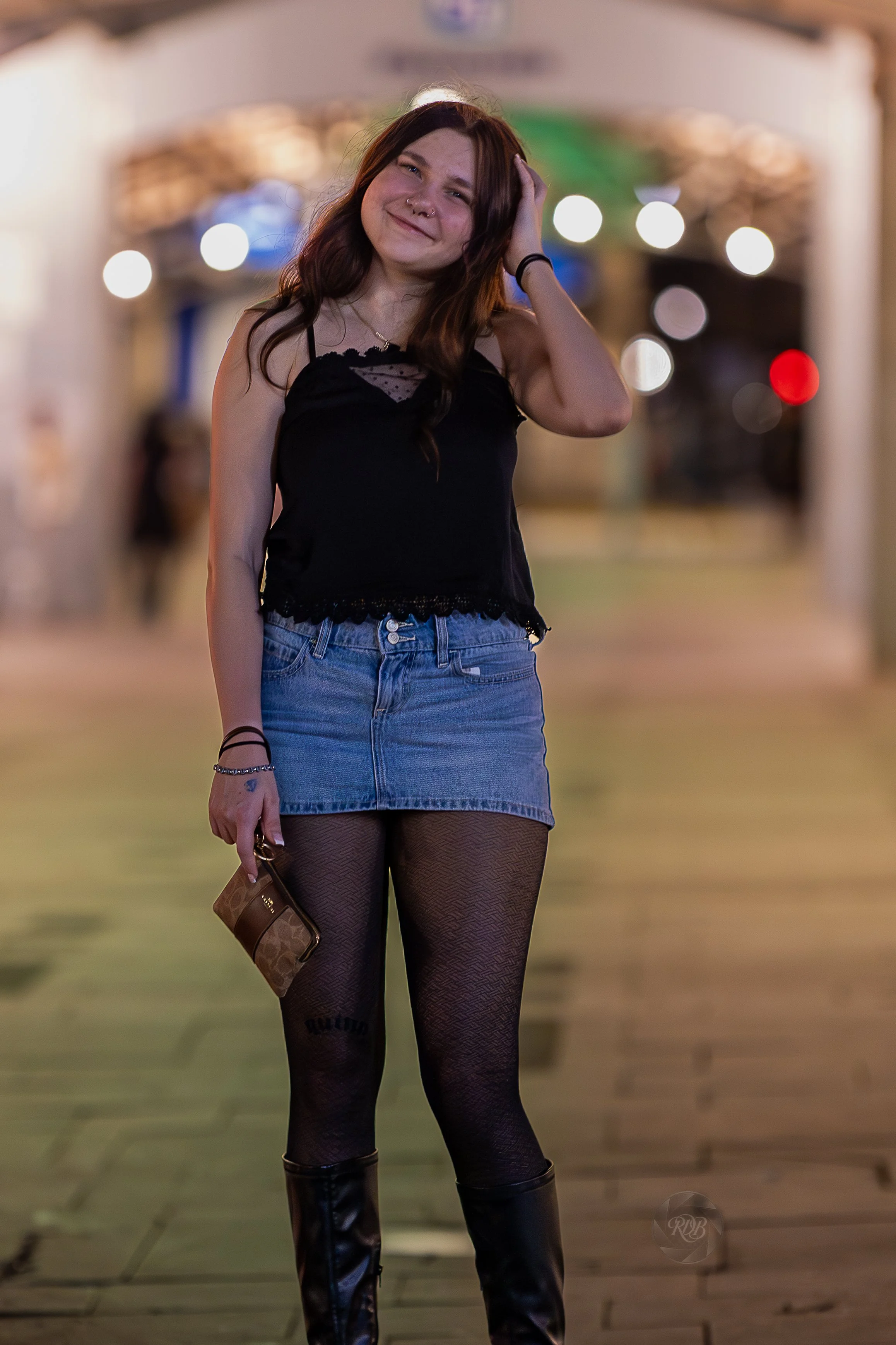 A young woman standing on a city sidewalk at night, smiling and touching her hair, wearing a black top, denim skirt, fishnet tights, and knee-high leather boots, with blurred city lights in the background.