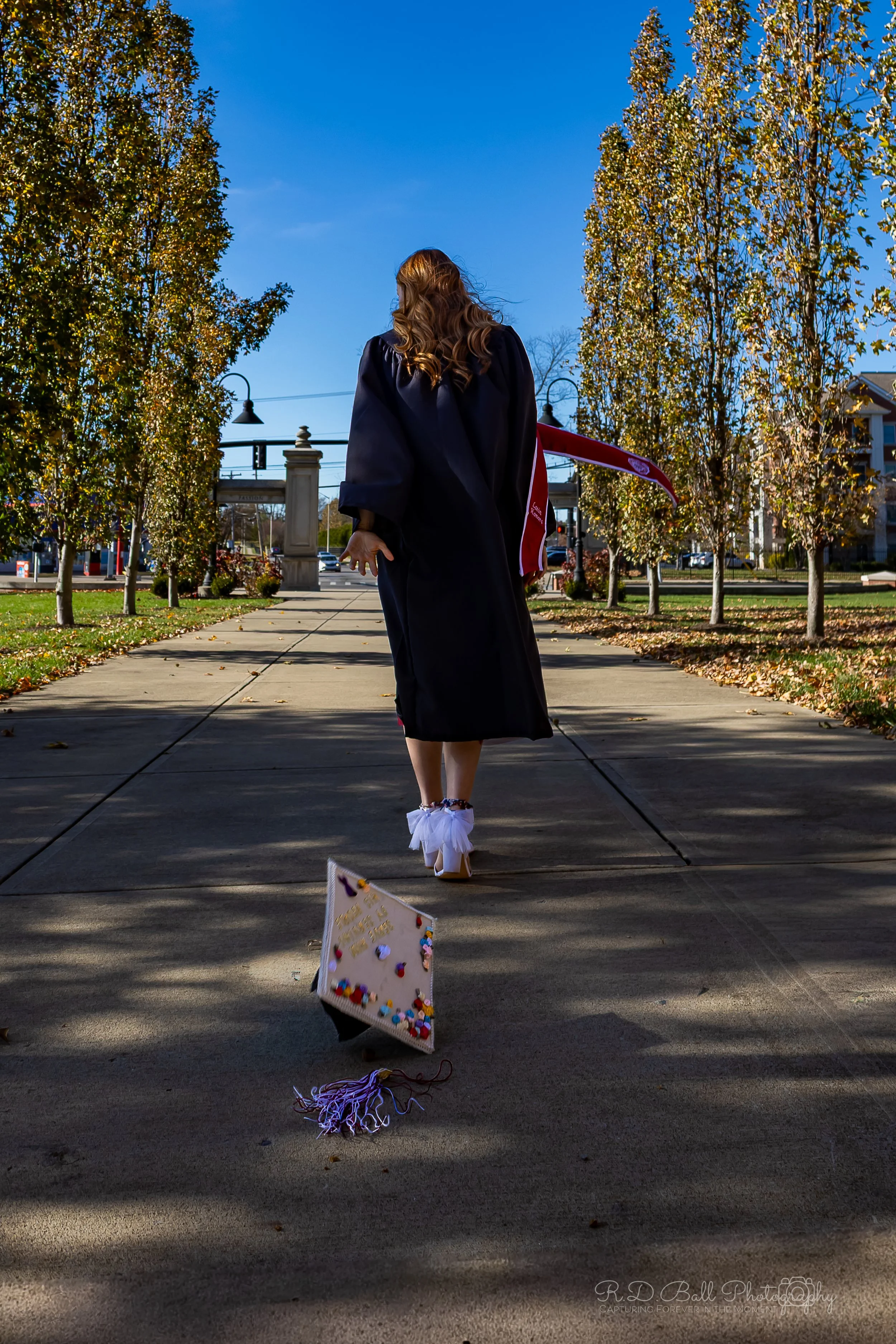 A woman in a graduation gown walking on a sidewalk during daytime, with a decorated graduation cap on the ground nearby, trees with fall foliage, and a blue sky in the background.