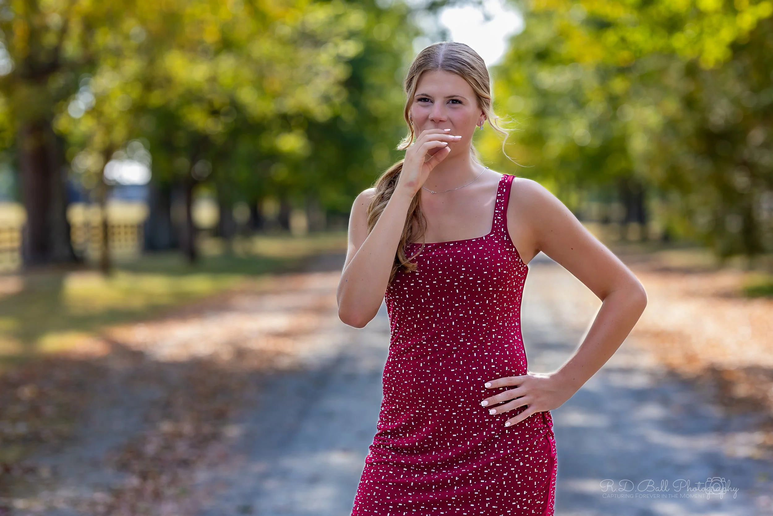 Young woman in a sleeveless red dress with white polka dots standing outdoors on a path surrounded by trees.