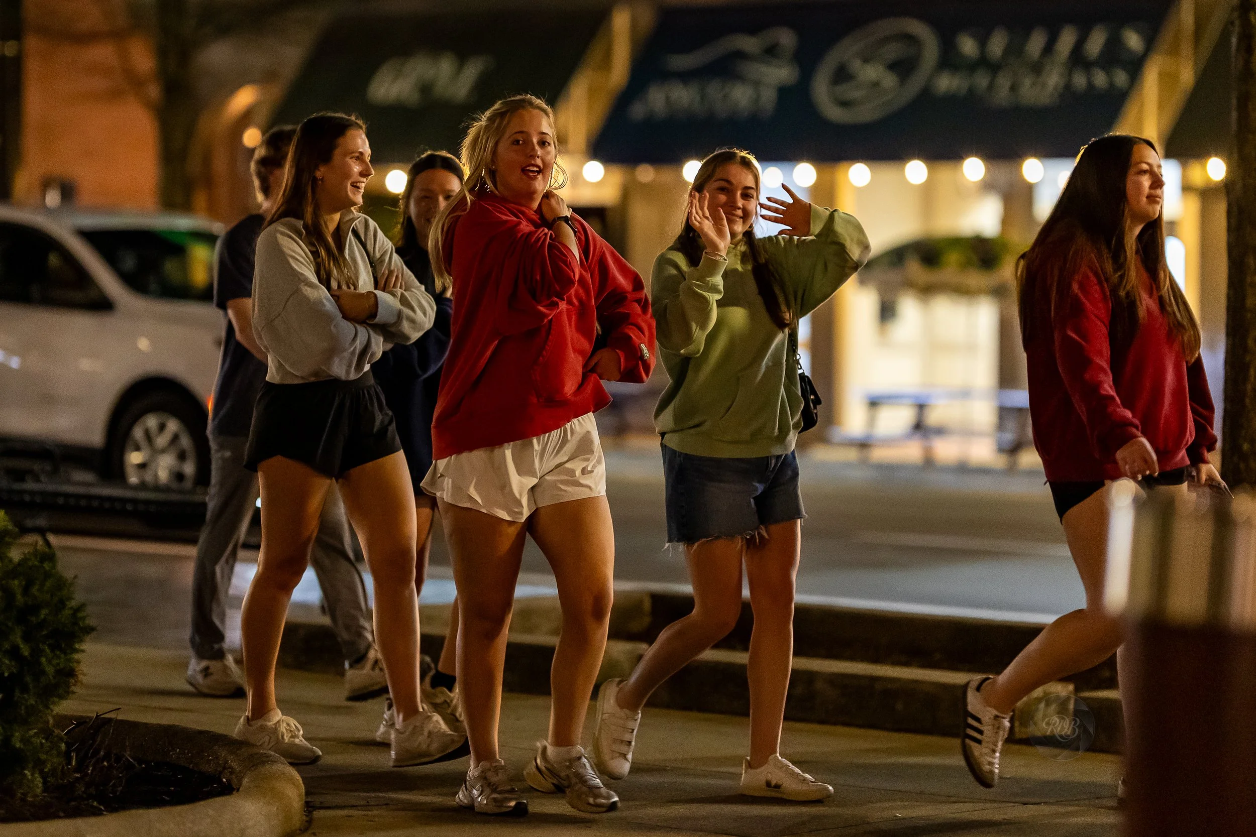 Group of young women walking at night on a city sidewalk, some smiling and talking, dressed in casual clothing and hoodies.