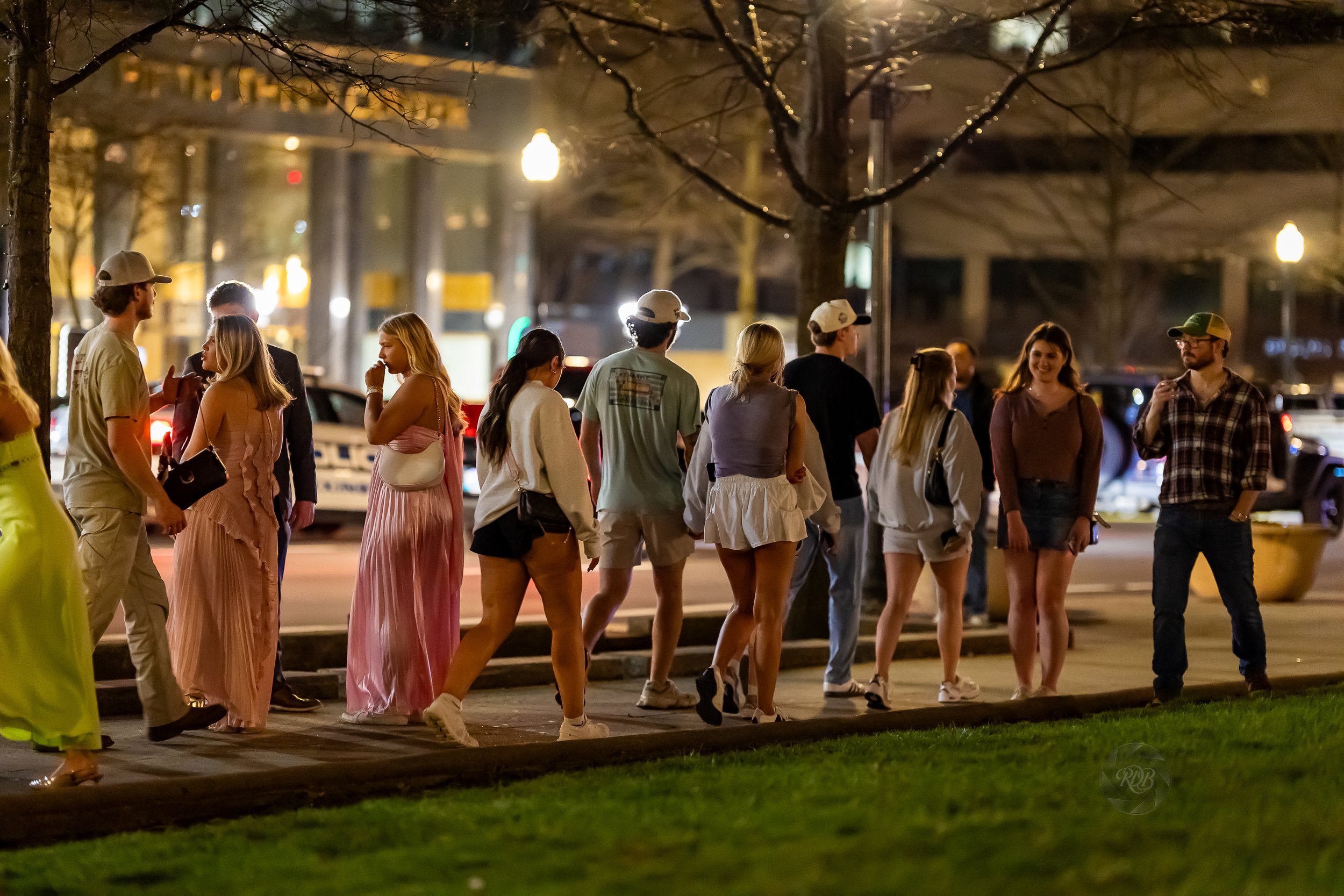 Group of people walking and talking on a city sidewalk at night, some in casual and some in stylish clothing, with cars and streetlights in the background.