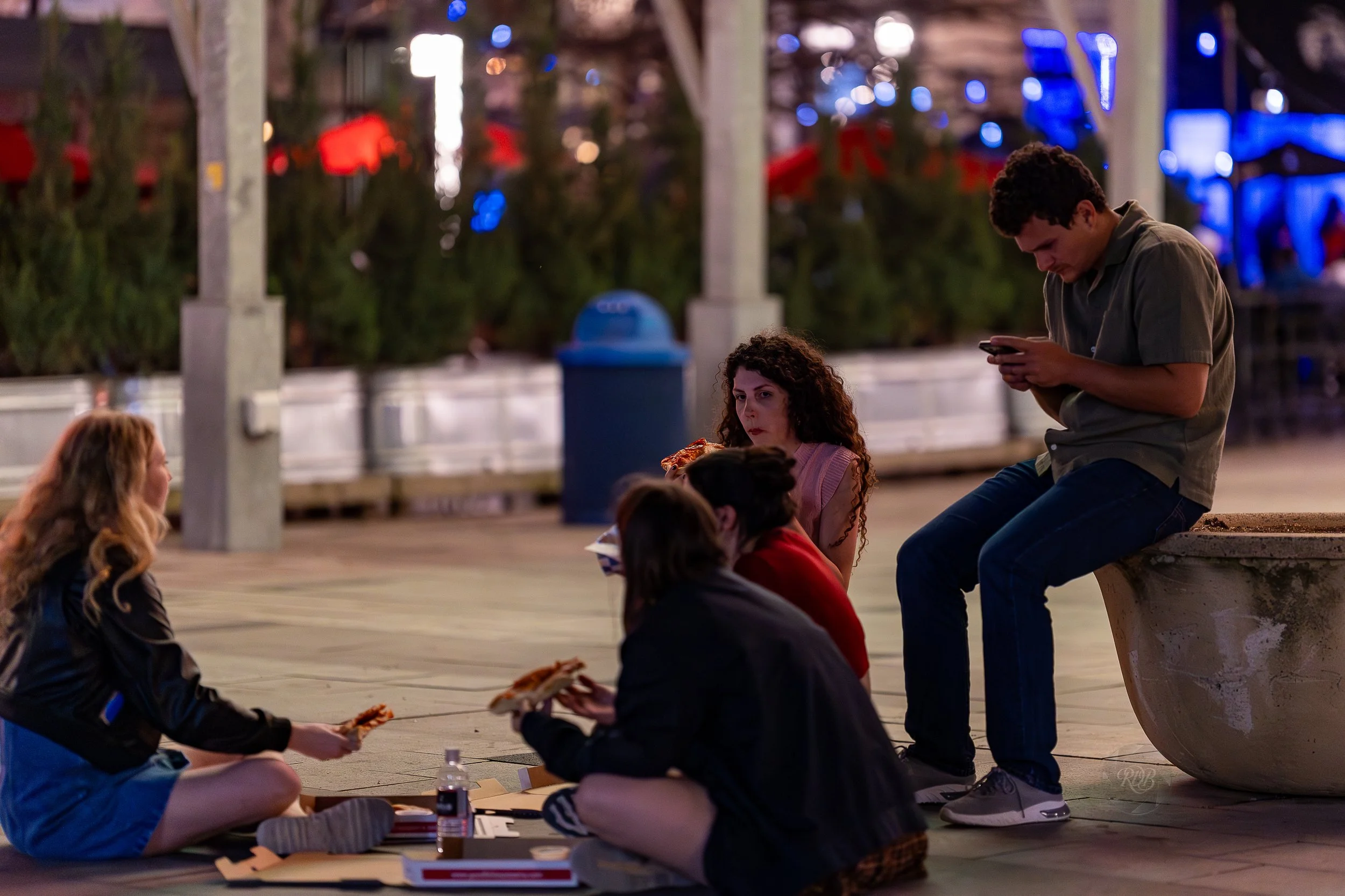A group of teenagers sits on the ground at an amusement park, eating pizza, with one standing girl looking at them. In the background, a boy is sitting on a concrete bench looking at his phone.