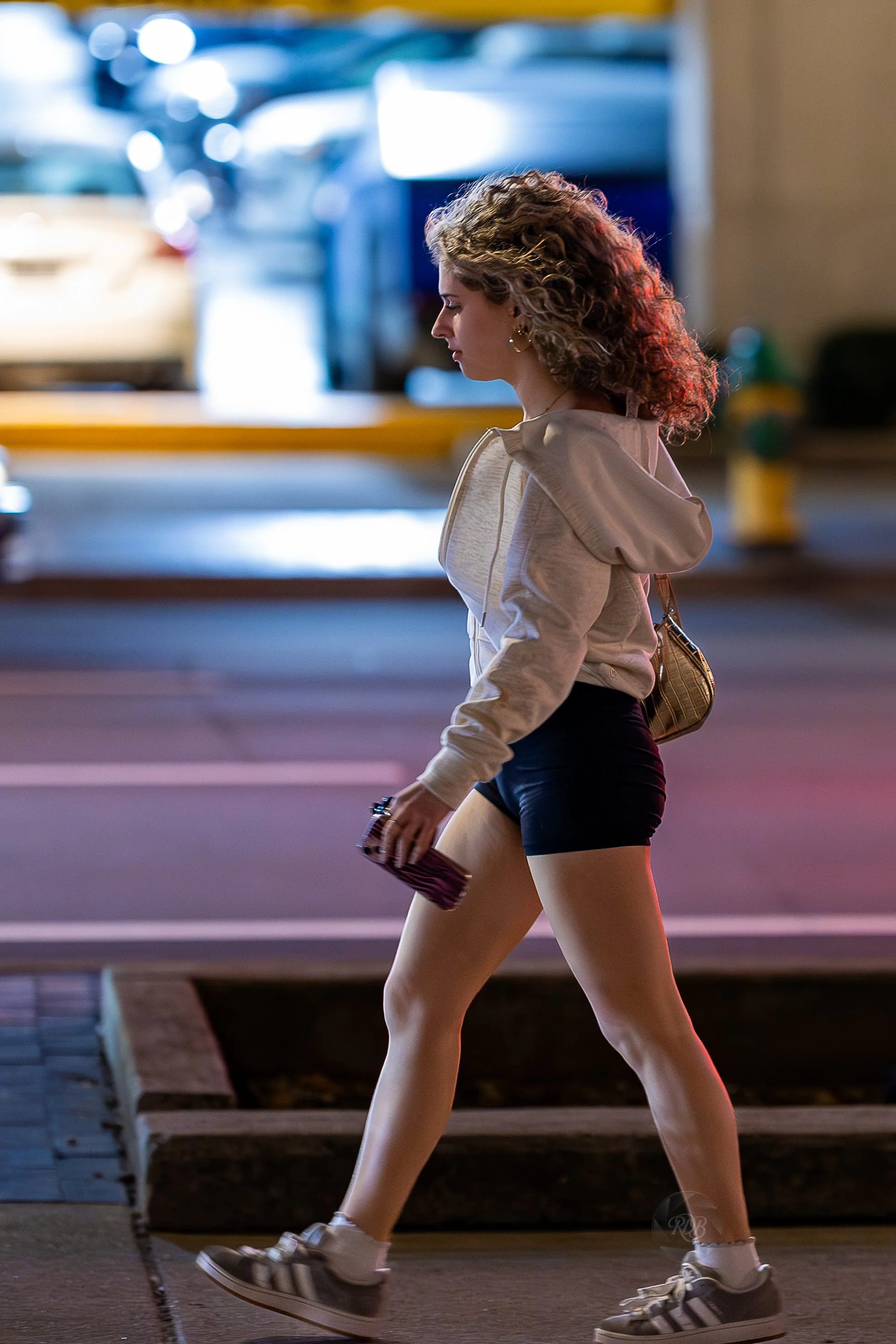 A young woman walking on a city sidewalk at night, wearing a beige hoodie, black shorts, and sneakers, carrying a small purse and holding a phone.