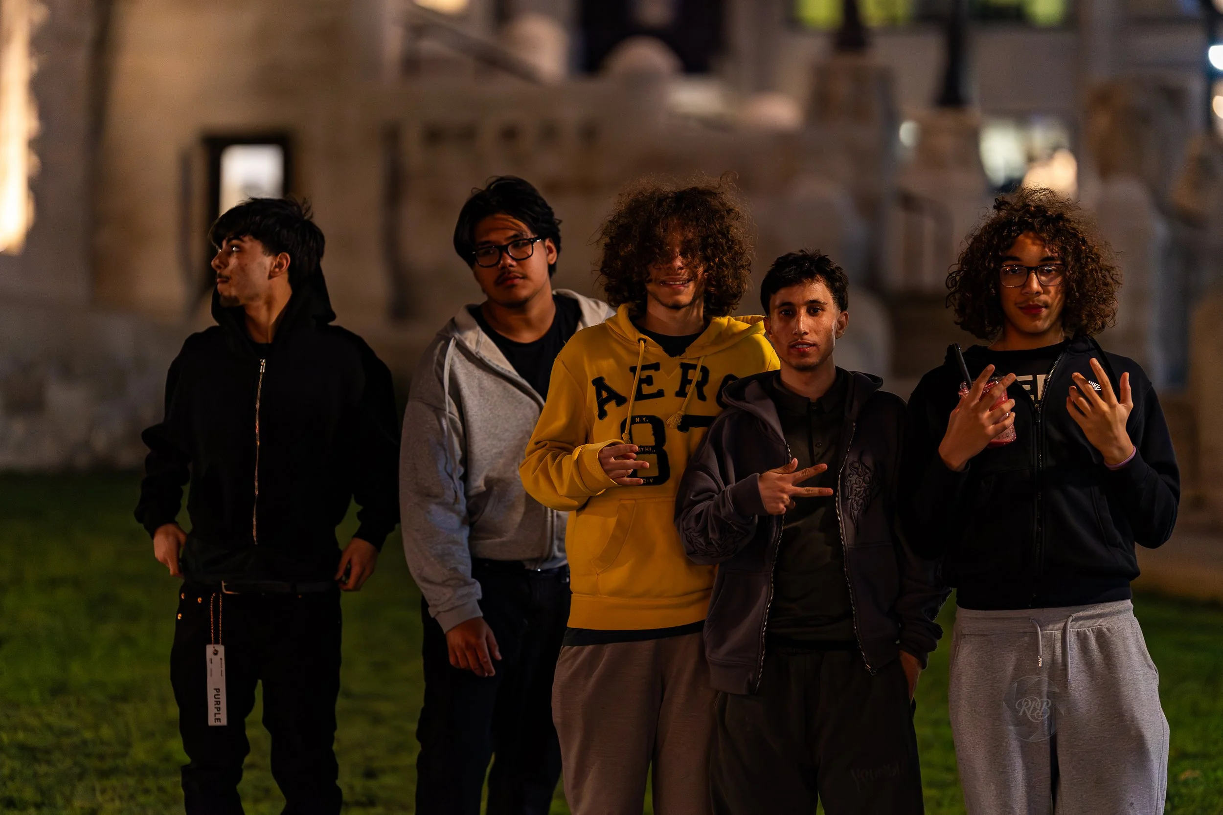 Five young men standing outdoors at night in front of a historic building made of stone, wearing casual hoodies and jackets, some making hand gestures, with a grassy area in the foreground.
