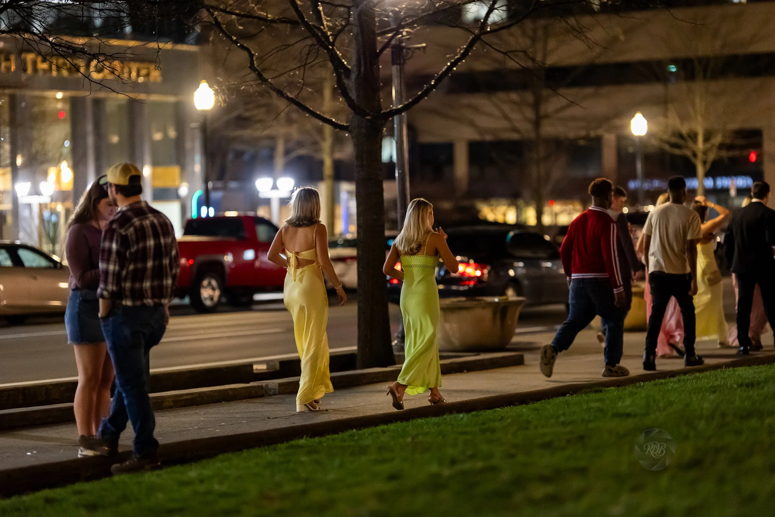 People walking on a city sidewalk at night, some wearing casual clothes and others in bright yellow dresses, with cars parked along the street and city lights in the background.