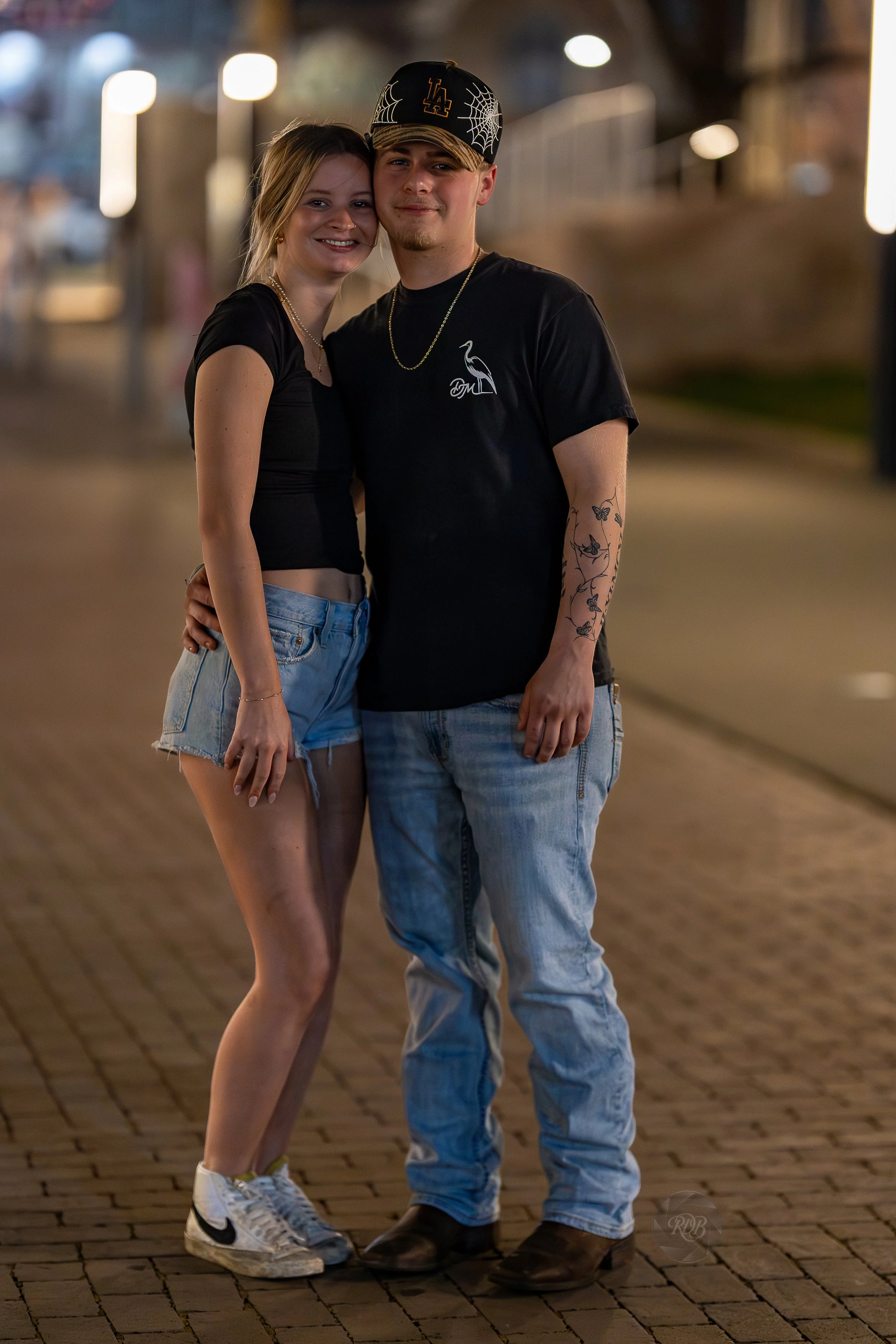 A young couple standing close together on a brick-paved street at night, smiling and embracing each other. The woman has blonde hair, wears a black crop top, denim shorts, and Nike sneakers. The man has tattoos on his arm, wears a black T-shirt, ligh