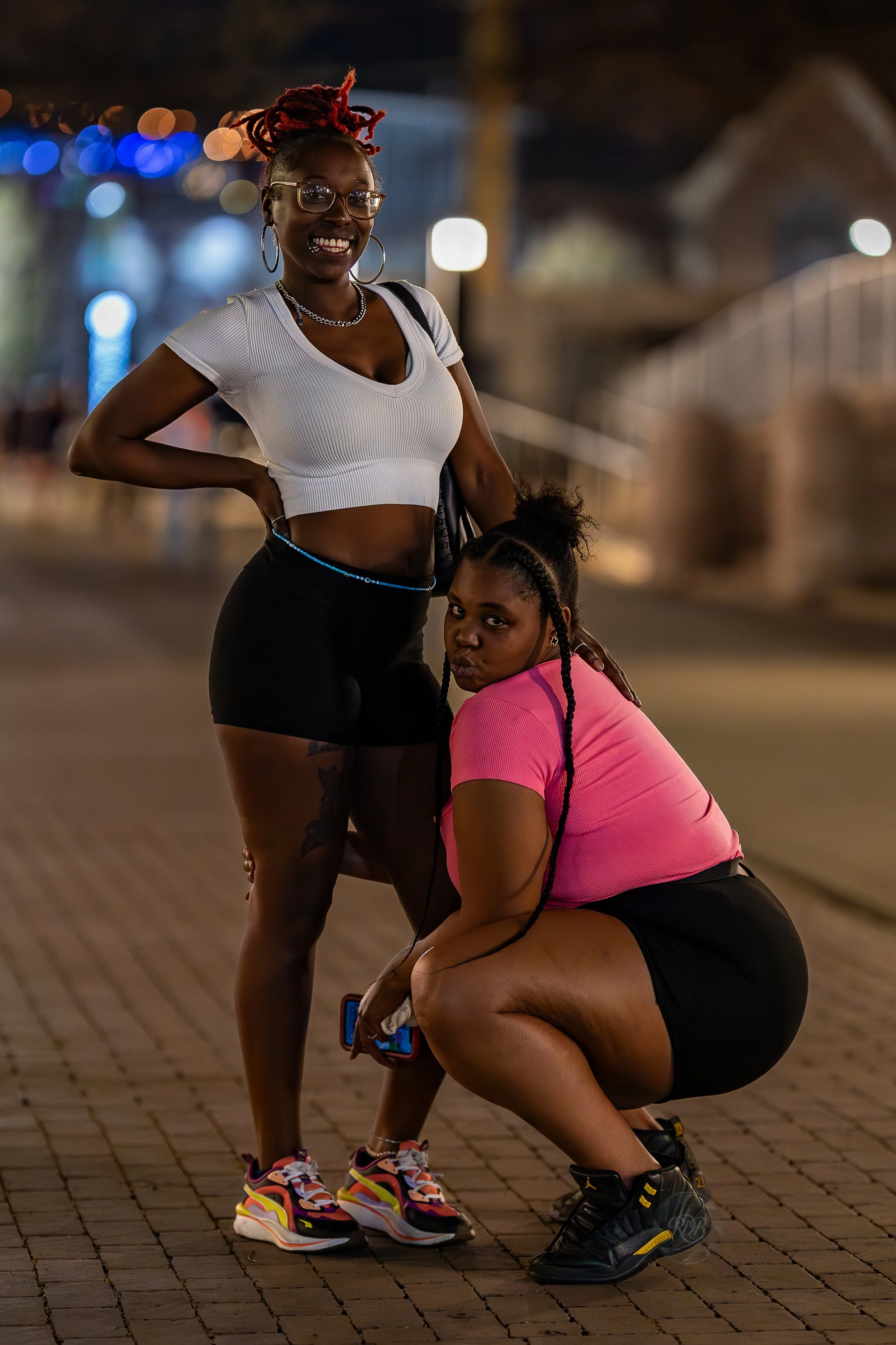 Two women in athletic clothing pose on a city sidewalk at night, with blurred colorful lights in the background.